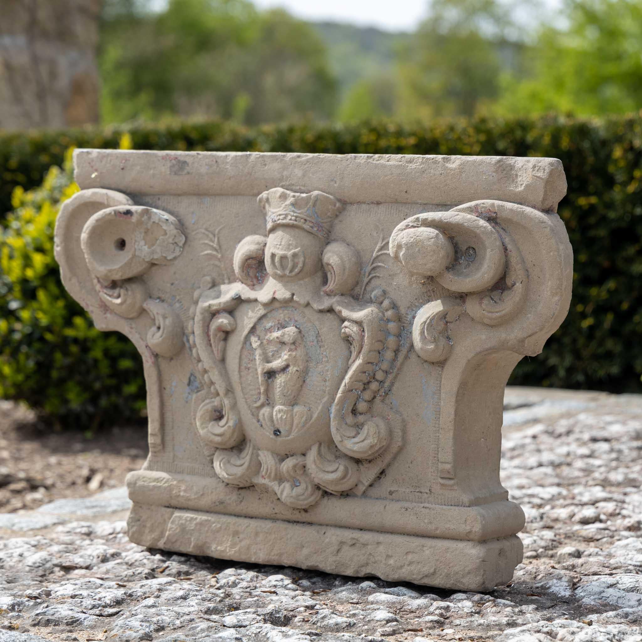 Large pilaster capital cut from sandstone with central coat of arms and volutes. Traces of old plaster.