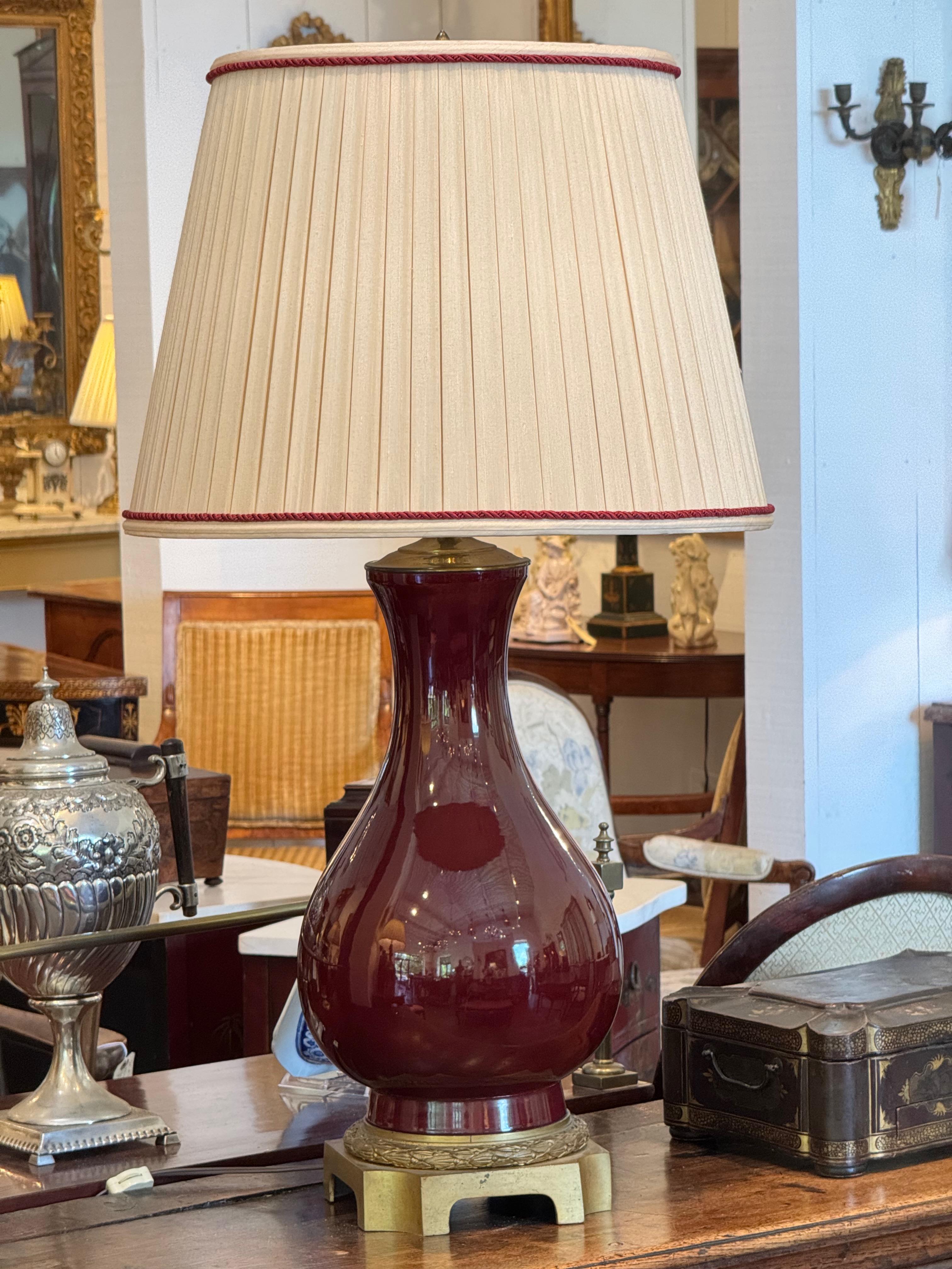 Elegant table lamp with a bulbous ceramic body in a deep sang de boeuf glaze, set on a brass footed base with laurel banding. Topped with a pleated cream shade trimmed in red, the lamp offers a classic silhouette with rich color and subtle detailing.