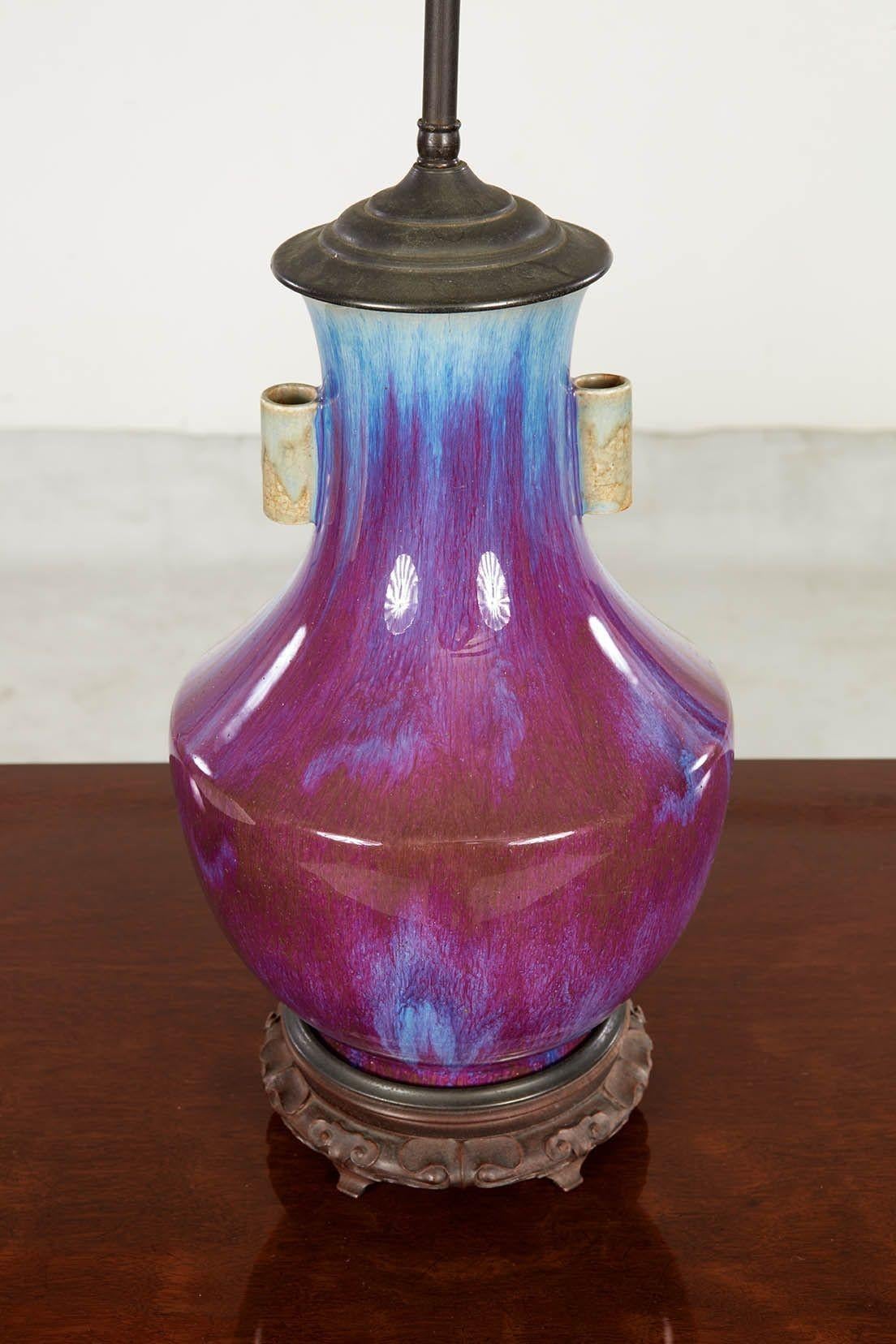 A Chinese export table lamp having vase shaped base with a lovely multi-hued sang de boeuf glaze sitting on a carved wooden vase stand and topped with a bronze cap with raised double sockets. China, circa 1910. Shade not included.