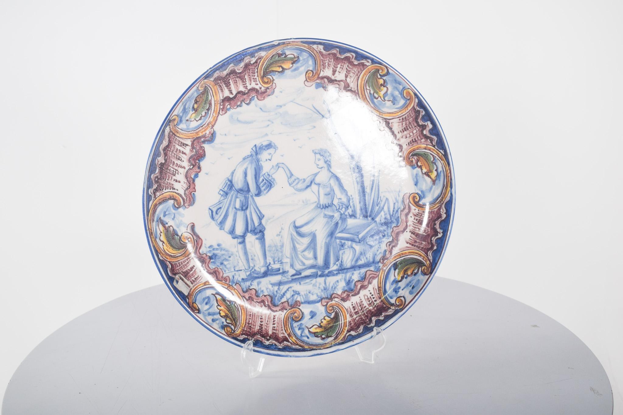 Sant'Anna Portuguese earthenware dish. Polychrome decoration, the centre depicting a romantic scene in which a gentleman bends down in a gallant gesture to kiss the hand of a lady sitting on a bench, in a bucolic and serene setting. The painting, in