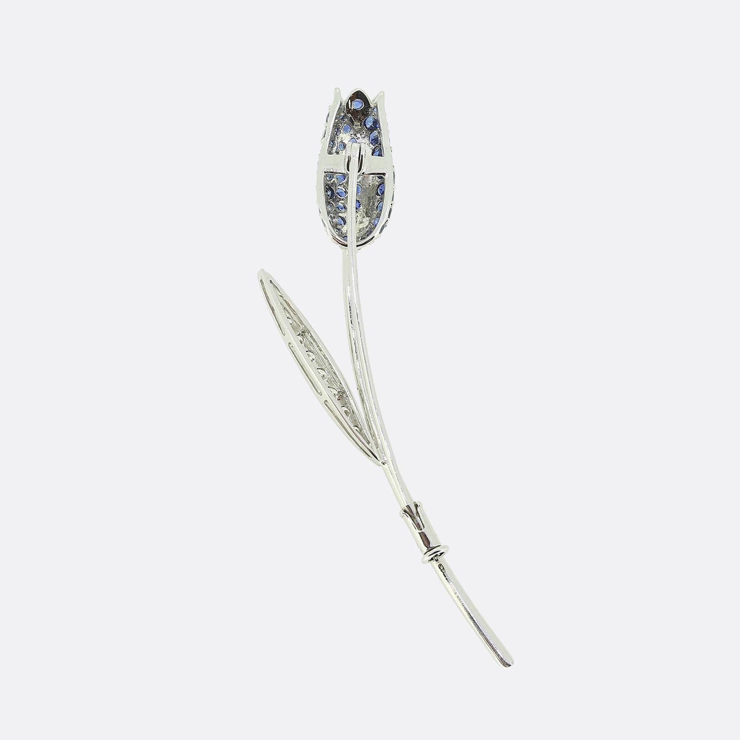 Here we have an elegant and refined brooch showcasing a tulip design. The flower is adorned with perfectly matched round faceted blue sapphires, set meticulously along the bud. A single leaf, encrusted with brilliant cut diamonds falls just below
