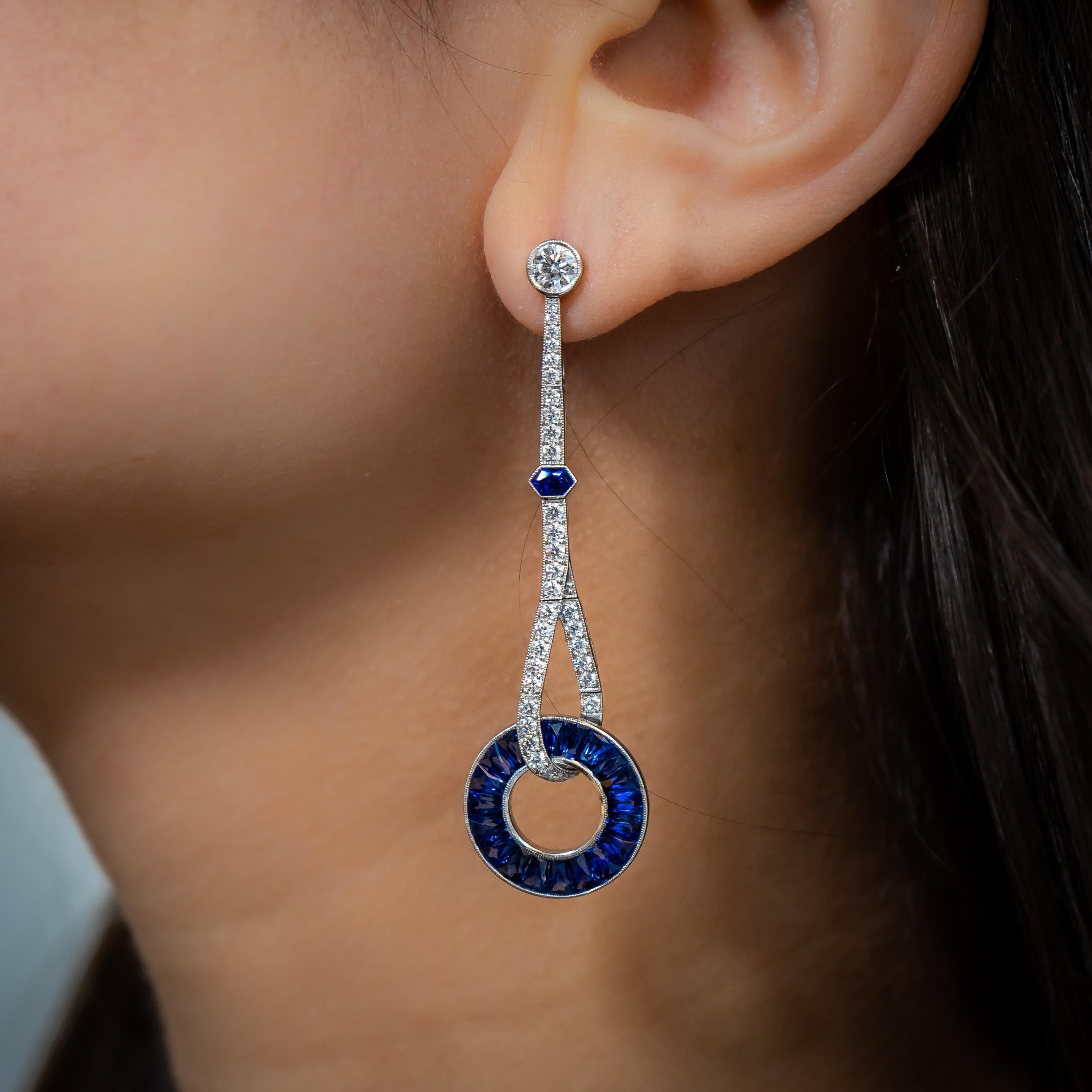 A pair of sapphire and diamond drop earrings, with diamond set tops, in rub over settings, with a total weight of 0.67ct, with pavé set drops, set with 66, round brilliant-cut diamonds, with a total weight of 0.92ct, each looping around a wheel of