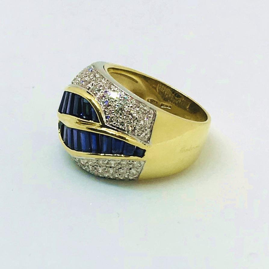 18K gold sapphire and diamond wide ring band centering two calibrated, channel-set “wave-like” rows of blue straight and tapered baguette-cut sapphires surrounded by pavé-set round brilliant cut diamonds. The ring measures approximately 16 mm wide