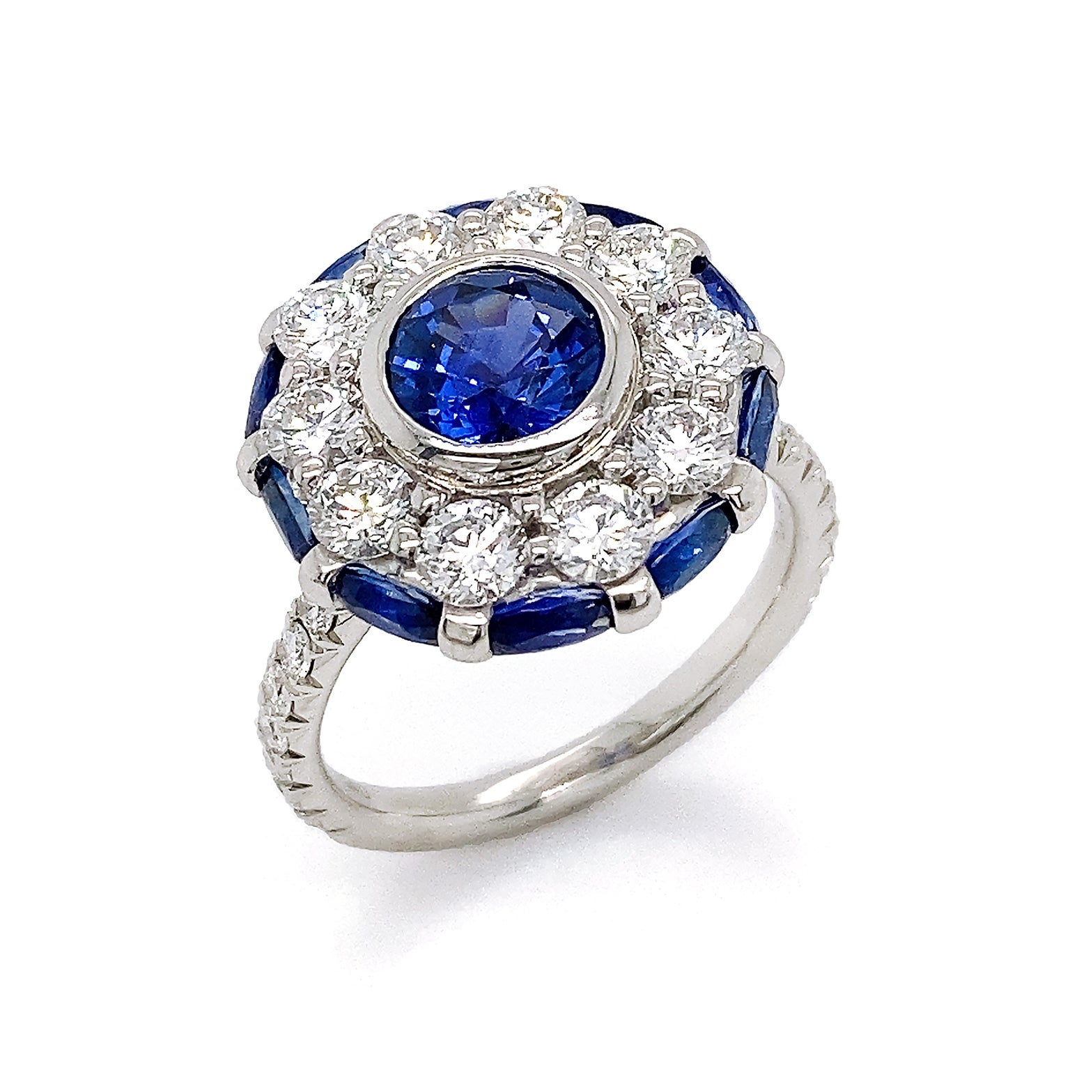 Iridescent blues and greens reflect from the center of this ring. A round sapphire shimmers as a halo of brilliant cut diamonds enclose it. Bordering the diamonds is an additional halo, made of oval sapphires. For both texture and light, brilliant