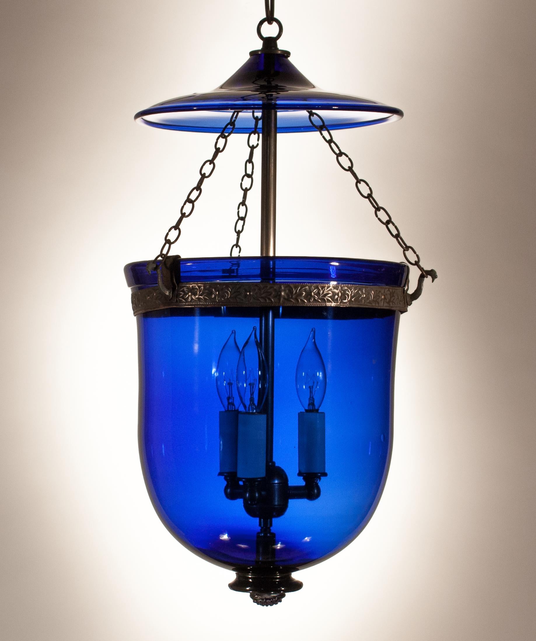 The brilliant sapphire blue of this circa 1880 English hand blown glass bell jar lantern makes it a true gem in our collection. The pendant has its original embossed brass band with floral decoration and glass smoke bell/lid. It has been newly