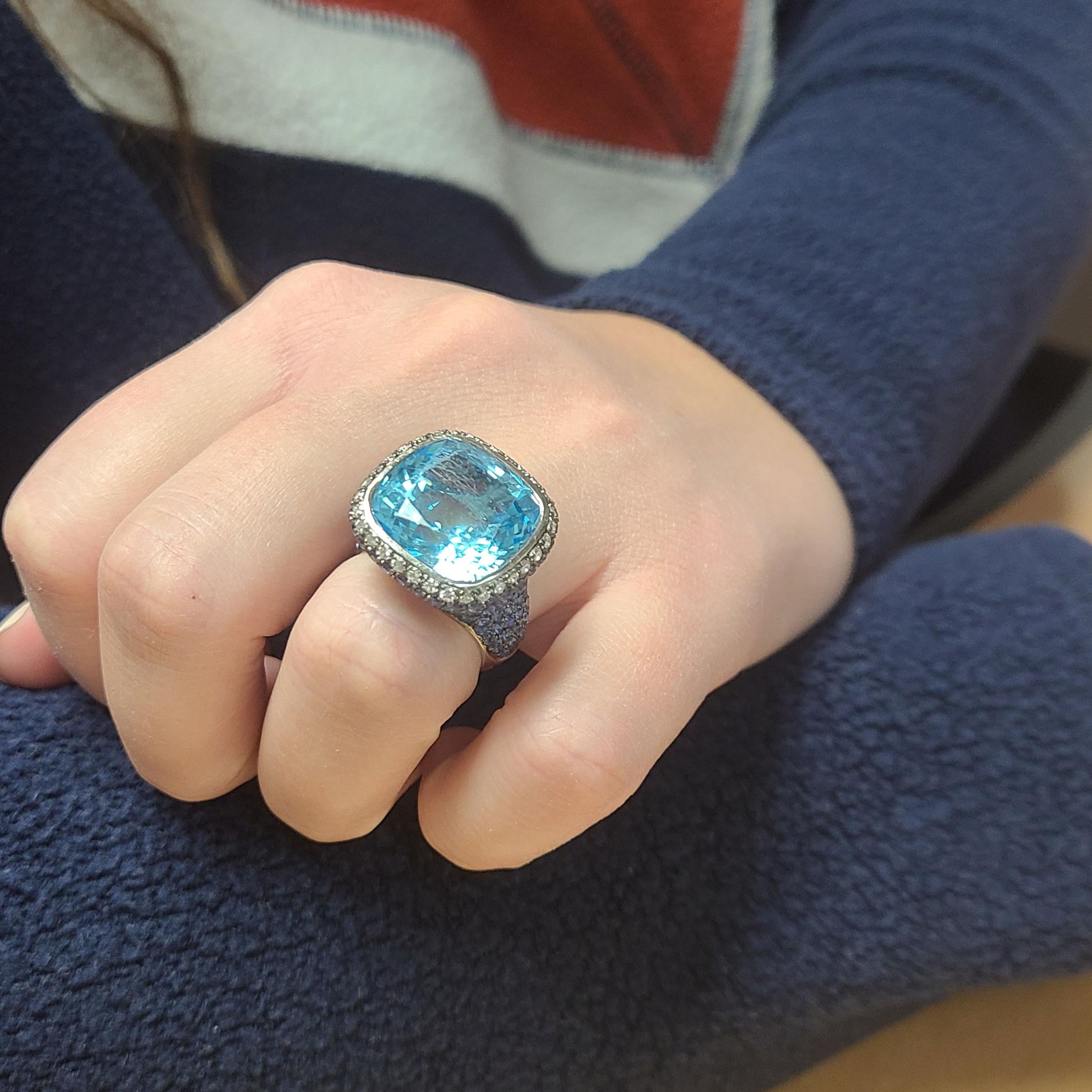 This ring is in 18K White Gold with a cushion cut blue Topaz at its center. It is surrounded by white Diamonds. Blue Sapphires run all the way down from the center down to the shank of the ring. The black background you see behind the Sapphires is