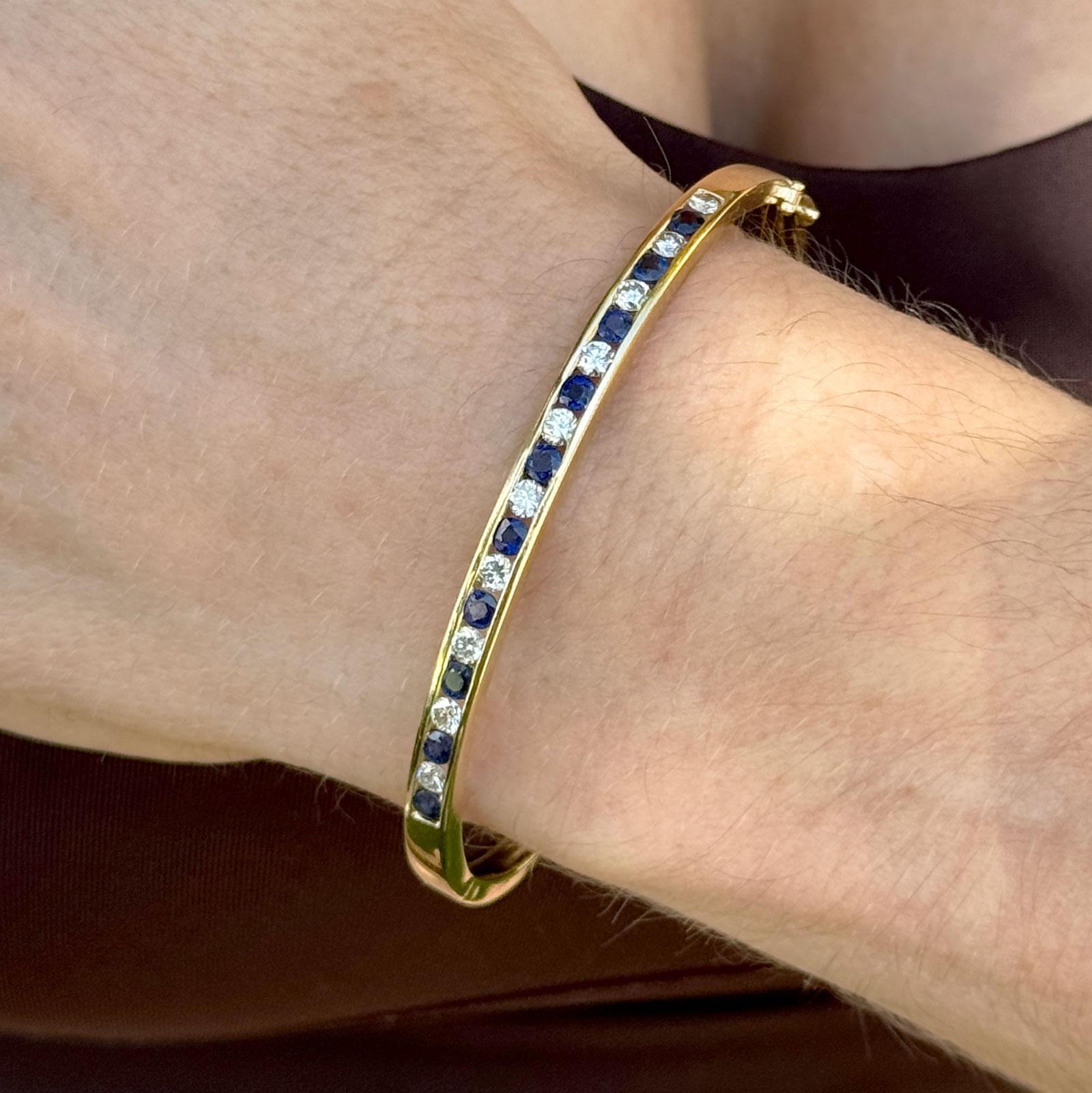 This elegant 18 karat yellow gold hinged oval bangle bracelet showcases a refined alternating design of 10 vibrant sapphires and 10 round brilliant-cut diamonds totaling approximately 1.00 carat, graded I-J in color and VS2 in clarity. Measuring 4mm