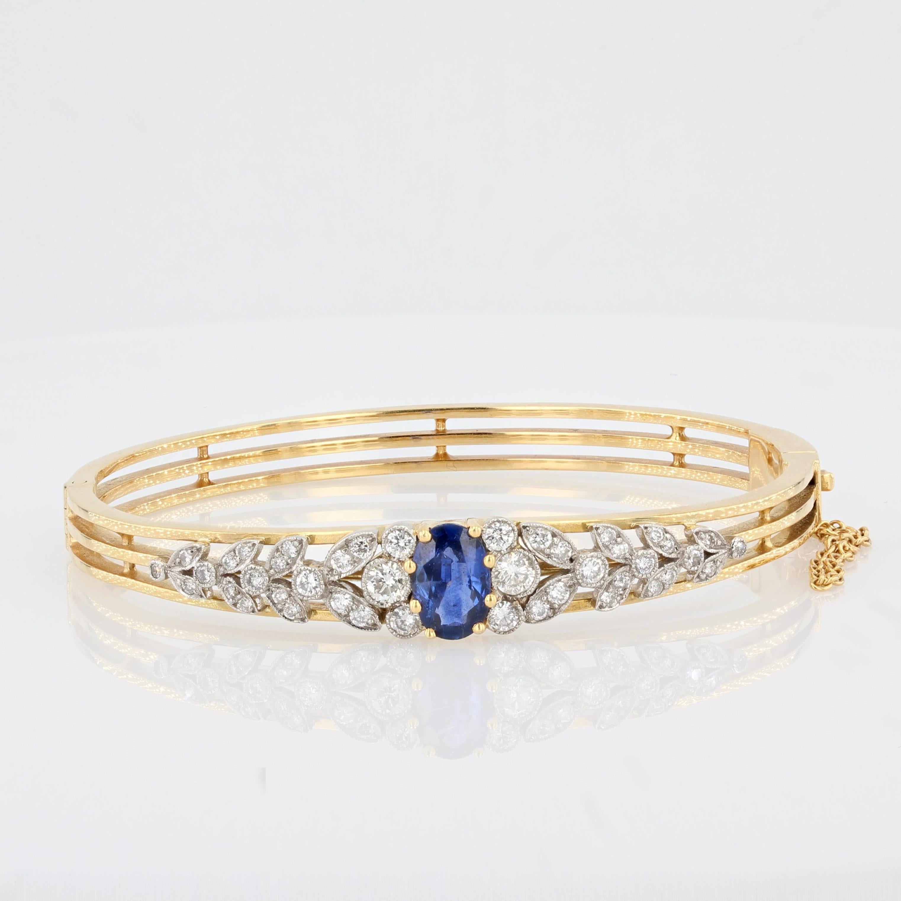 Bracelet in 18 carat yellow gold, eagle head hallmark. 
This superb opening oval bangle bracelet features 3 flat gold strands with a floral design at the front entirely bezel set with brilliant cut round diamonds and a central claw set oval blue