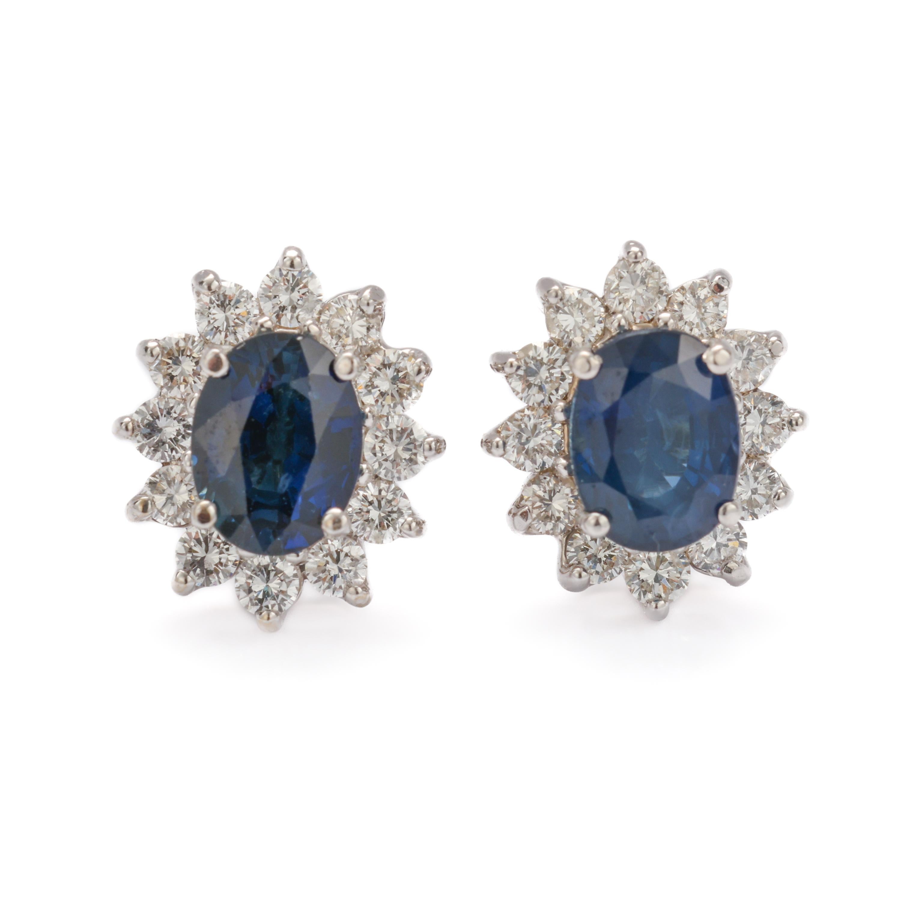 This classic pair of natural blue sapphire and diamond earrings was created in the 1990s -circa 1997- in 18K white gold. Each earring features an approximately 8mm x 6mm oval sapphire weighing slightly over one carat. Each sapphire is encircled by a