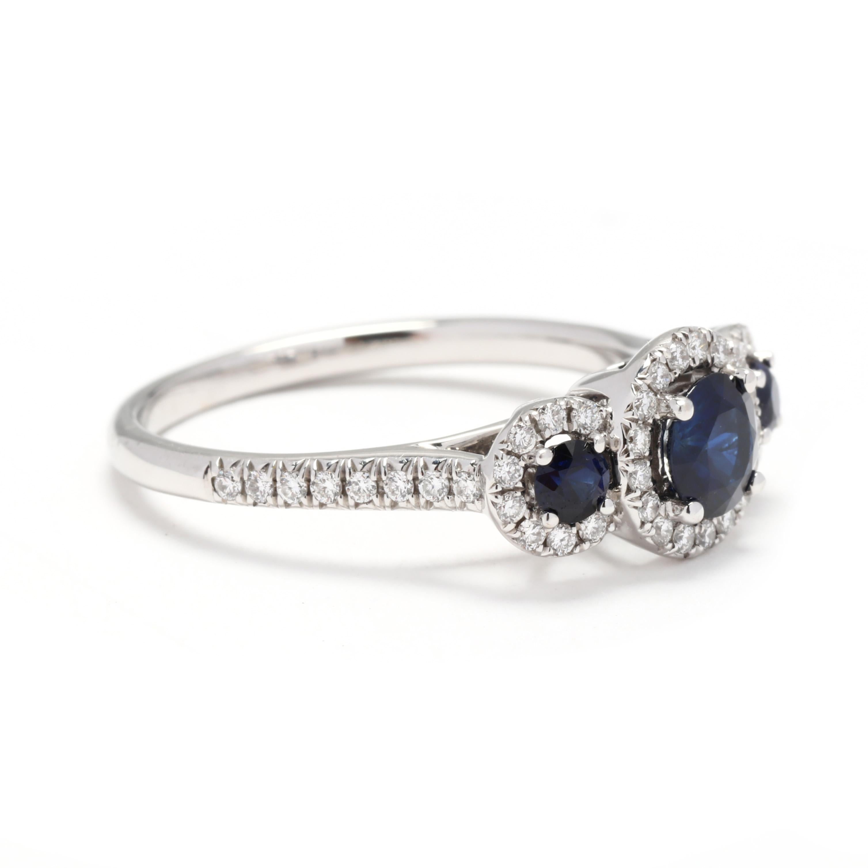 A 14 karat white gold natural sapphire and diamond three stone engagement ring. This ring features three round cut natural blue sapphires weighing approximately 1 total carat, each with a halo of round brilliant cut diamonds and diamonds down the