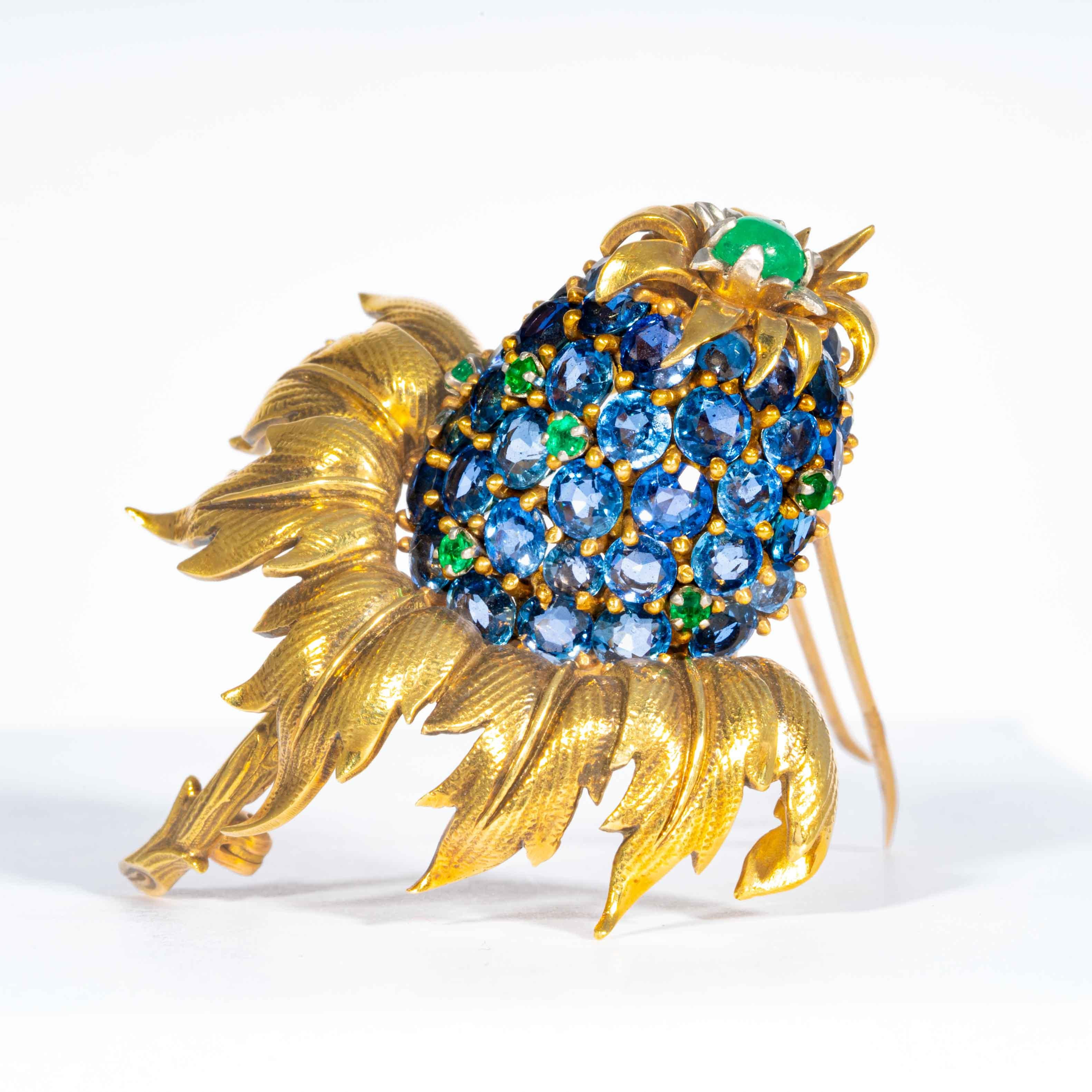 This late 20th century pin is comprised of full cut blue sapphire, and green emeralds set in 18kt yellow gold in this whimsically classic floral thistle themed design. This pin measures approximately 1.5