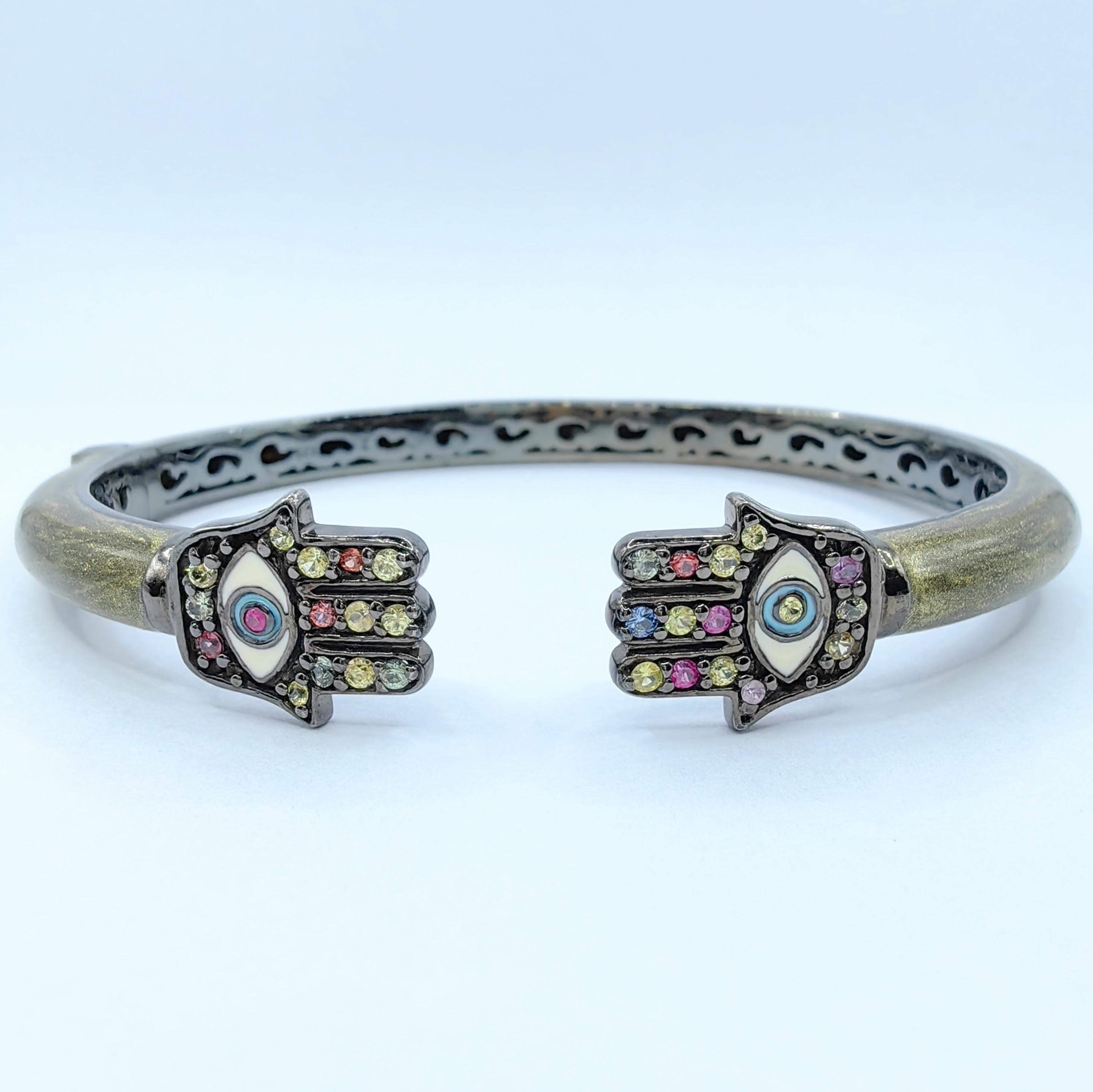 Dieses unverwechselbare, mit Saphiren besetzte Hamsa-Handschellenarmband verbindet meisterhaft uralte Schutzsymbole mit modernem Flair. Das Armband zeigt zwei Hamsahände, die mit leuchtenden, mehrfarbigen Saphiren von insgesamt 0,59 Karat verziert