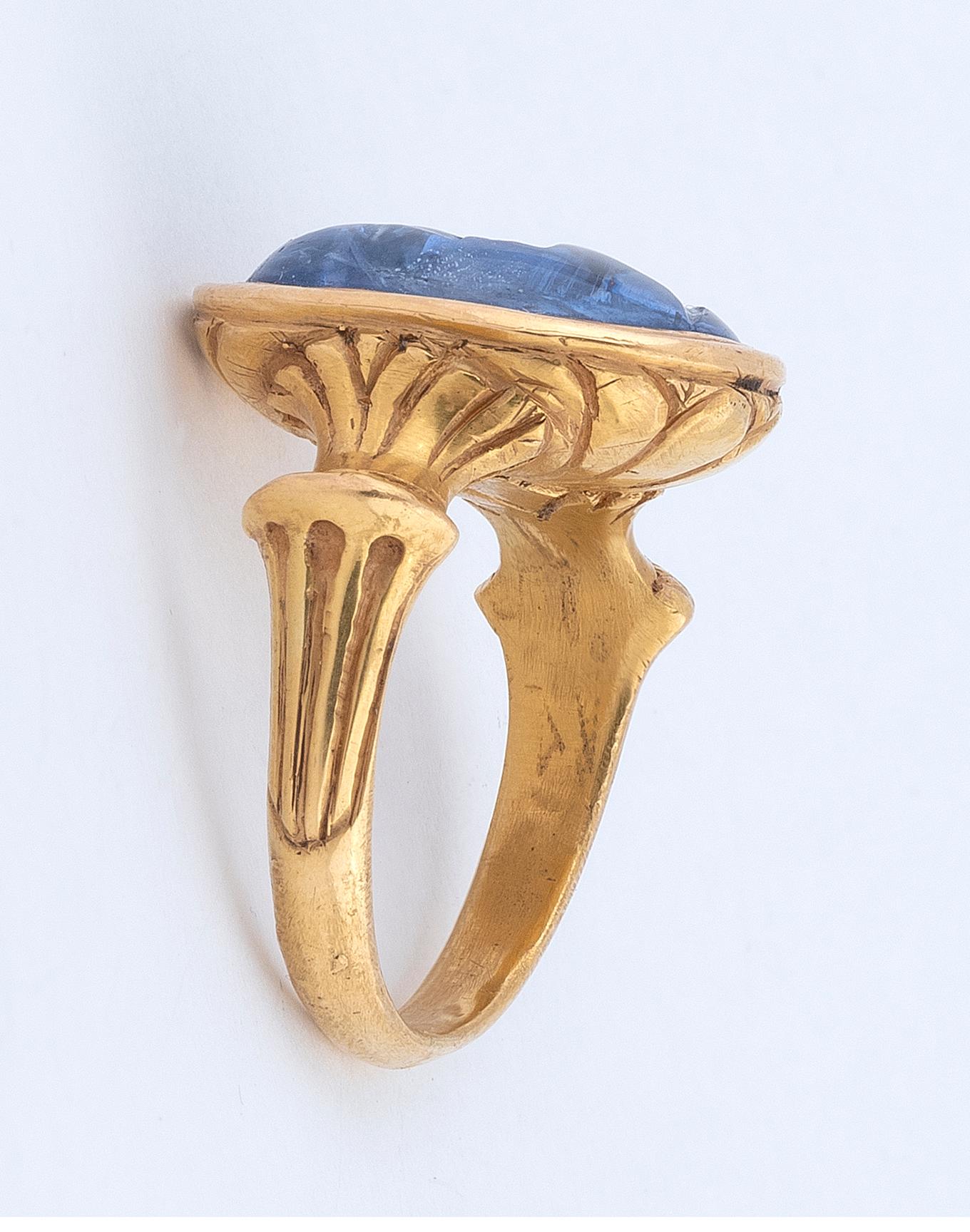The oval-shaped sapphire carved to depict the profile of a Roman Emperor Caligola, in a closed-back setting, between foliate shoulders, 19th century composite, ring size 9 1/2
Weight : 21,20gr.
Size of bezel : 14mm x 18mm