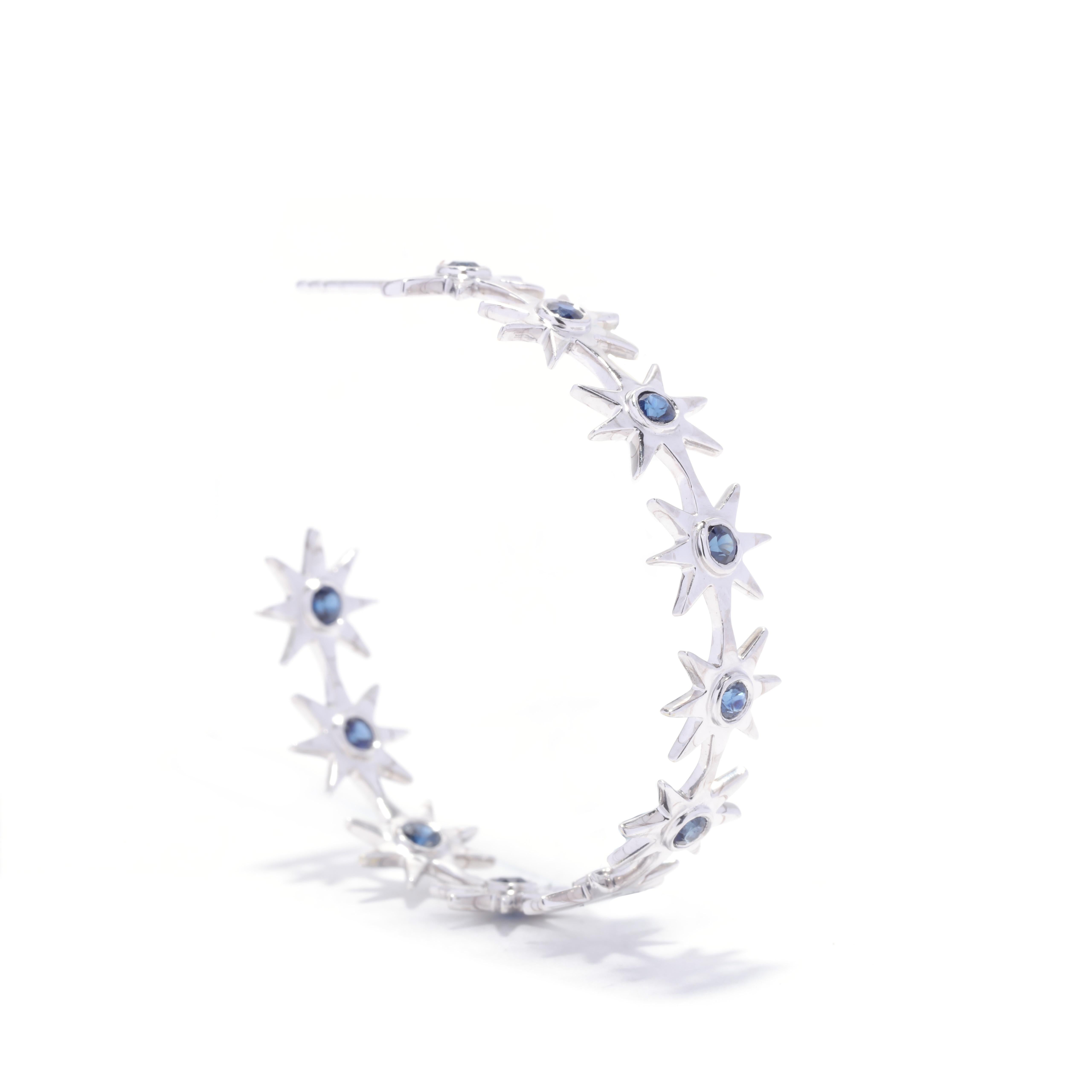 A pair of 18 karat white gold sapphire star hoop earrings. These large funky hoops feature a repeating pattern of star shapes, each with a bezel set round cut sapphire weighing approximately 2 total carats and with pierced push backs.

Stones:
-