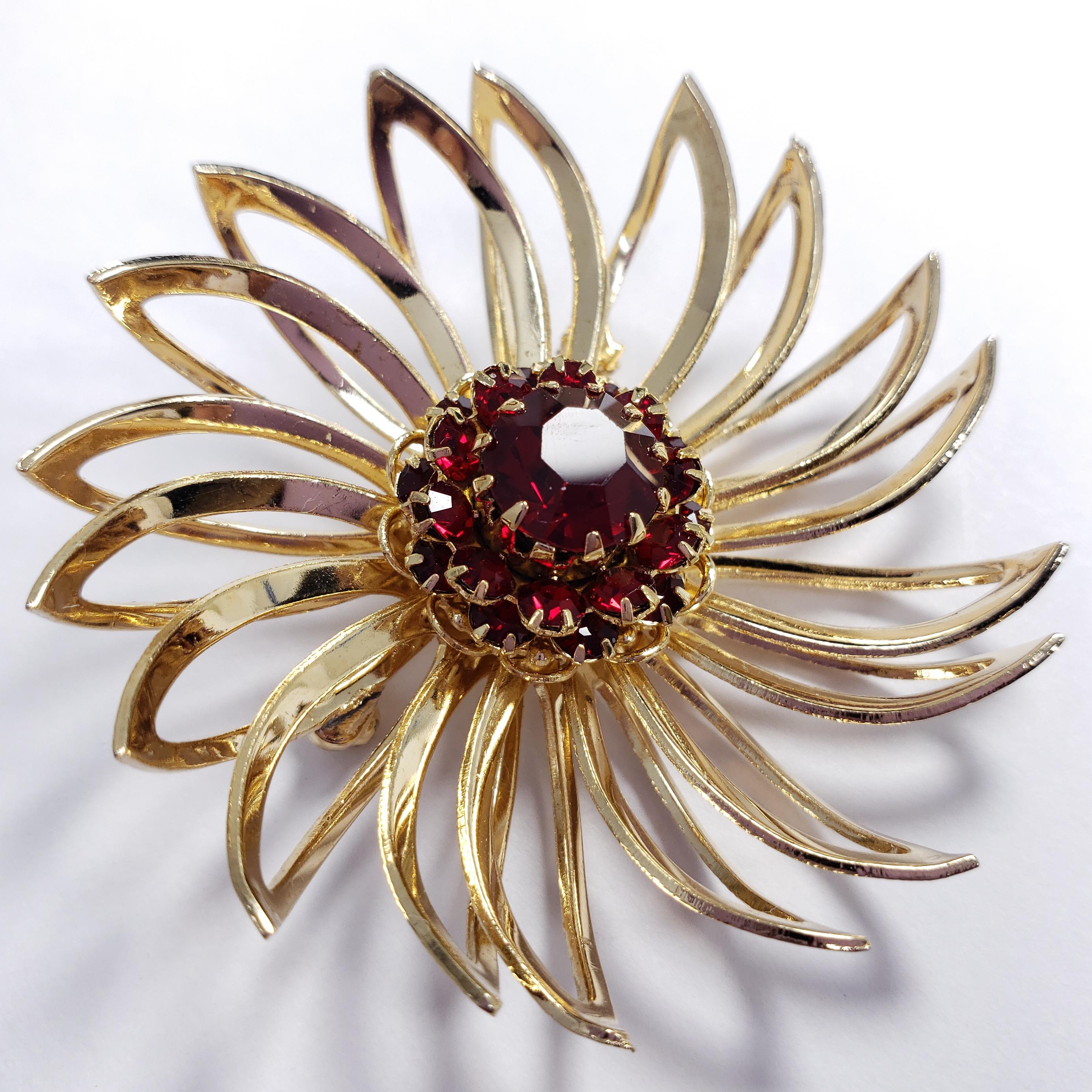 A stylish pin brooch by Sarah Coventry. Features curved goldtone petals and a centerpiece faceted red crystal, accented with smaller crystals.

Hallmarks: © SARAH COV