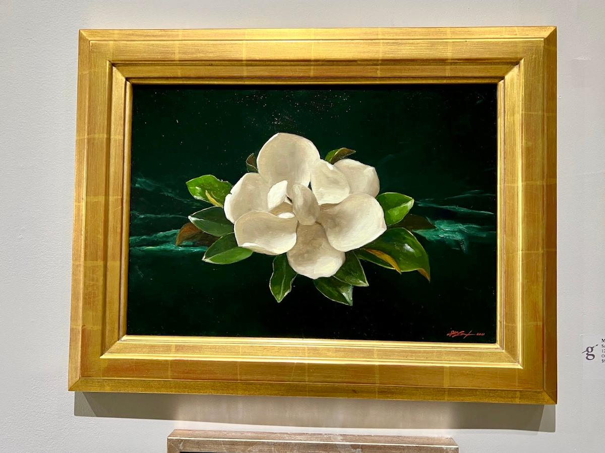 A single magnolia flower blooms out of a background of green-tinted darkness.

Framed dimensions: 17.5 x 22.5 inches

Artist Bio
Sarah Lamb is a talented and dynamic realist painter. With classical skill—and through transparency, depth and