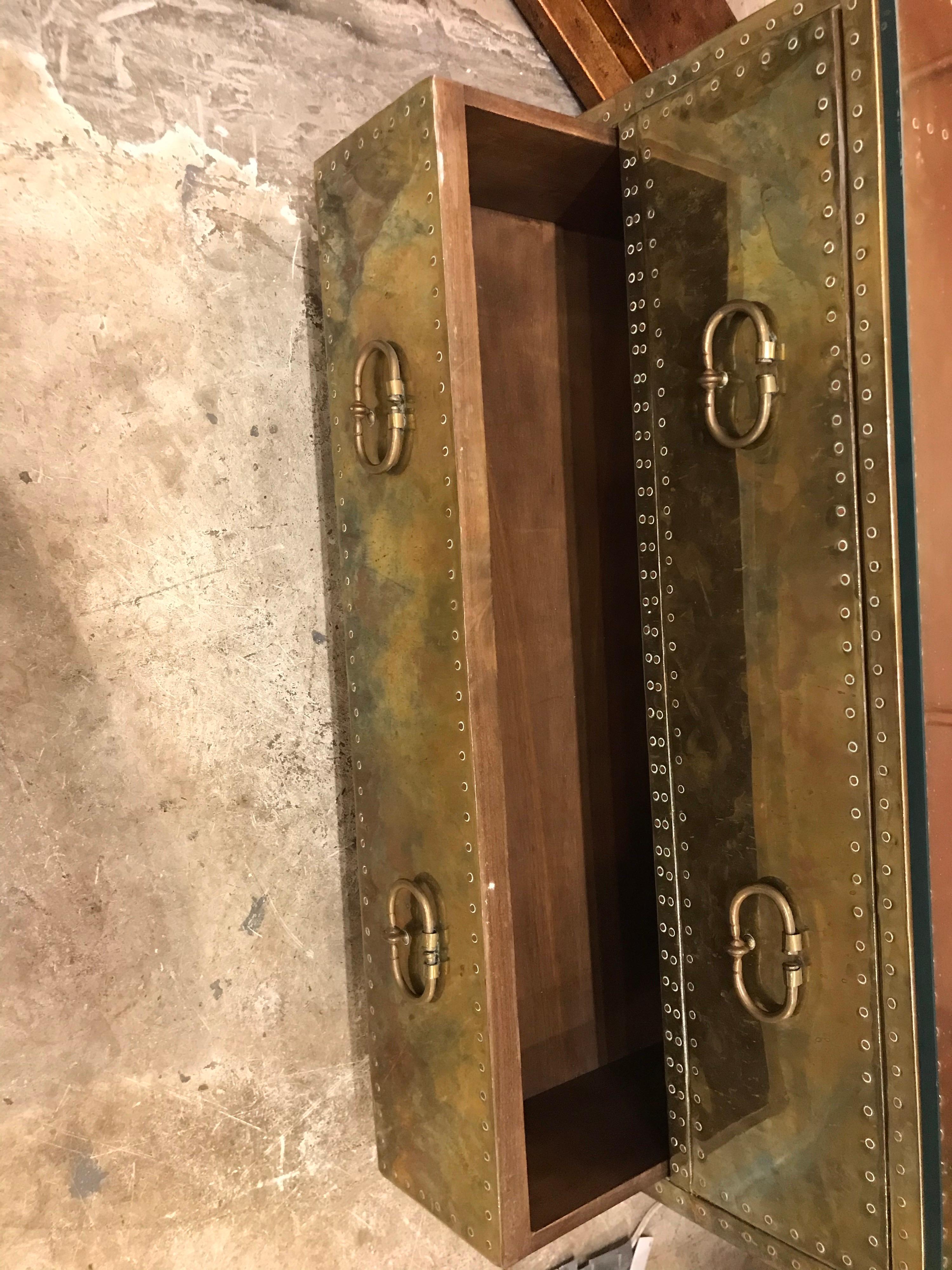 This is a brass clad two-drawer chest or trunk with copper nail heads and bun feet. It is made by Sarreid probably in the 1970s-1980s and has a glass top to protect it.