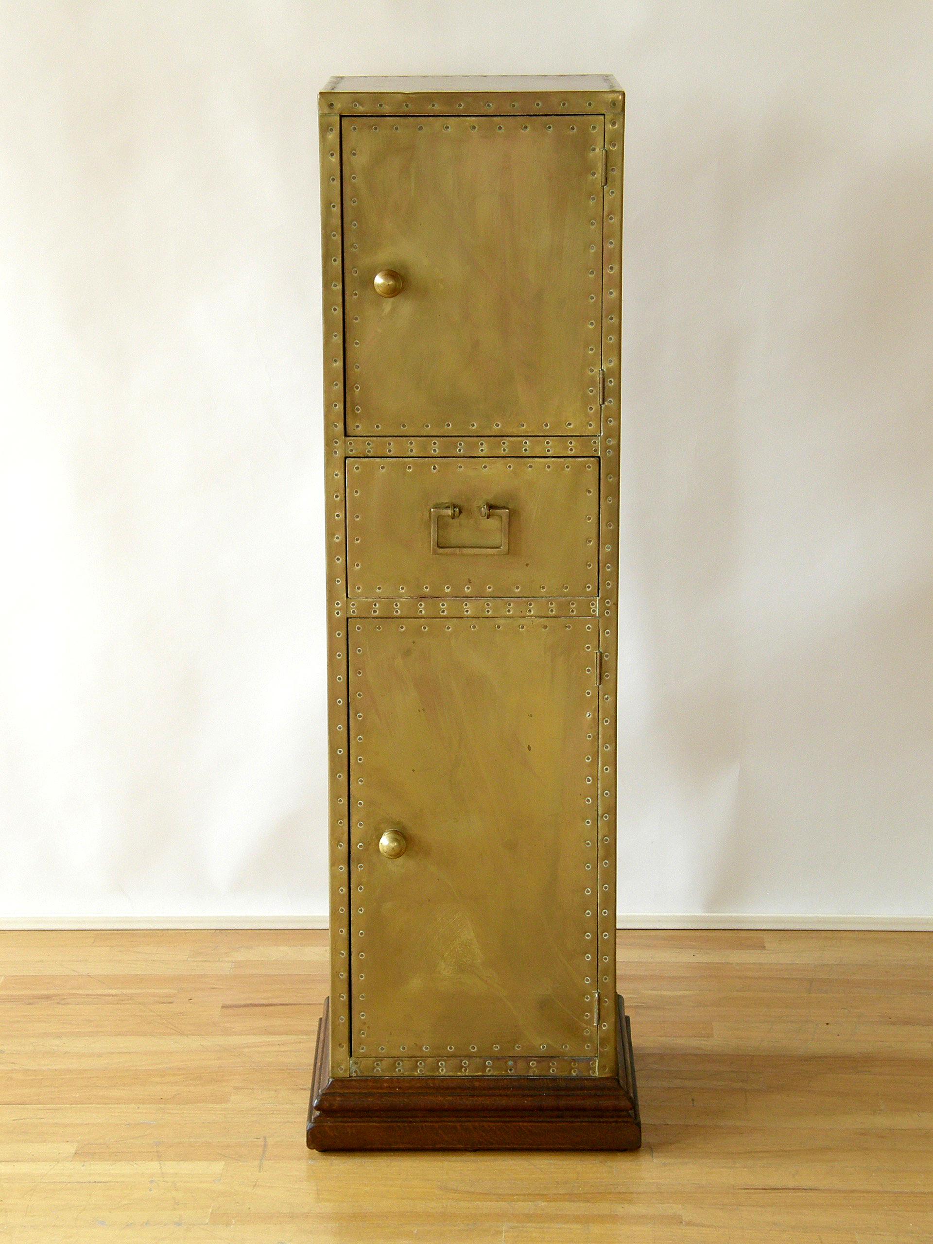 This tall, slender, brass-clad cabinet by Sarreid is shaped like a pedestal with storage. The nailheads are both functional and decorative as they create linear patterns that highlight the form. There is a smaller door on top and a larger door on