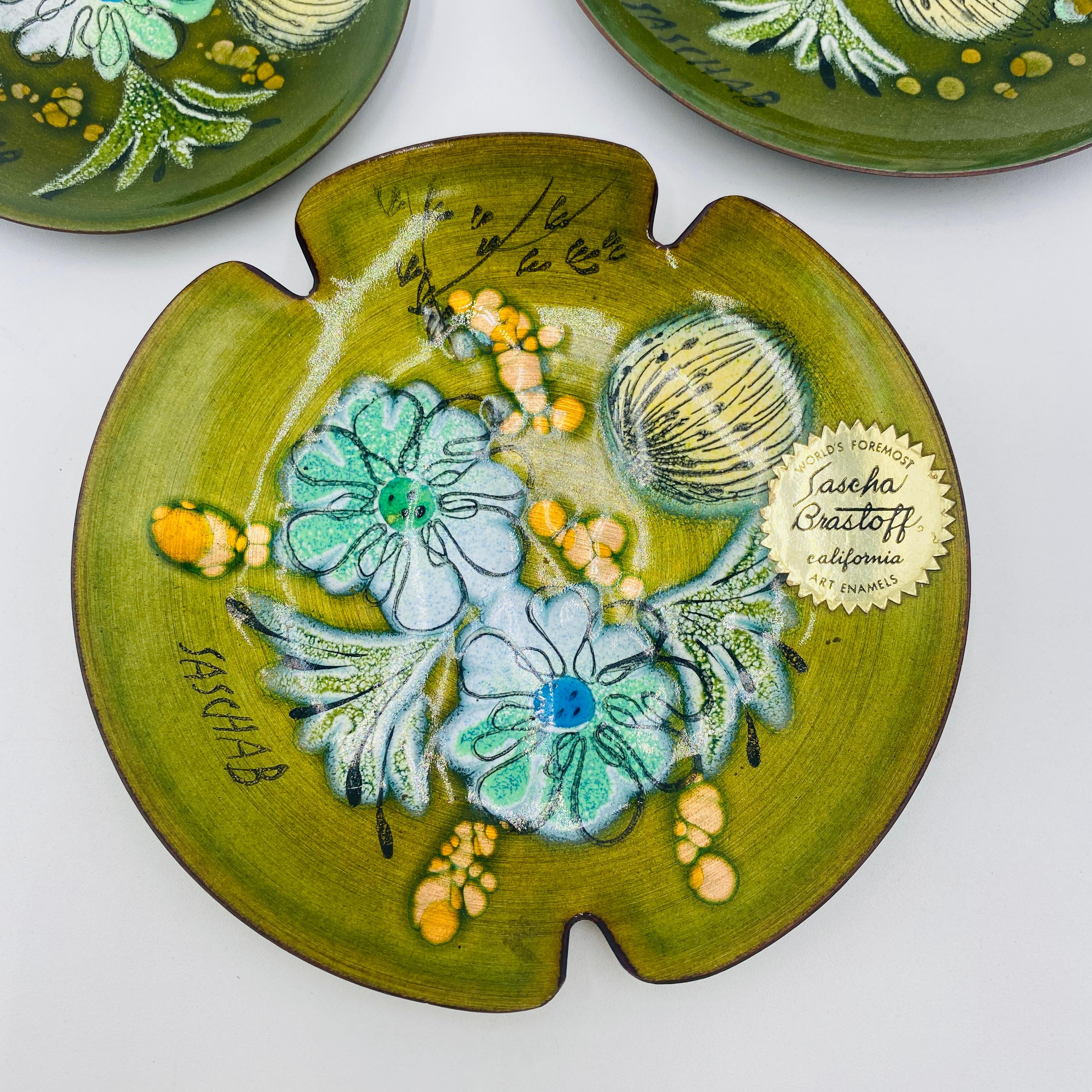 A vintage set of 3 mid-century green porcelain enamel copper ashtray dishes by Sascha Brastoff. The set features porcelain enamel over copper of gorgeous floral designs. The signature is in the interior of each ashtray and one of them retains the