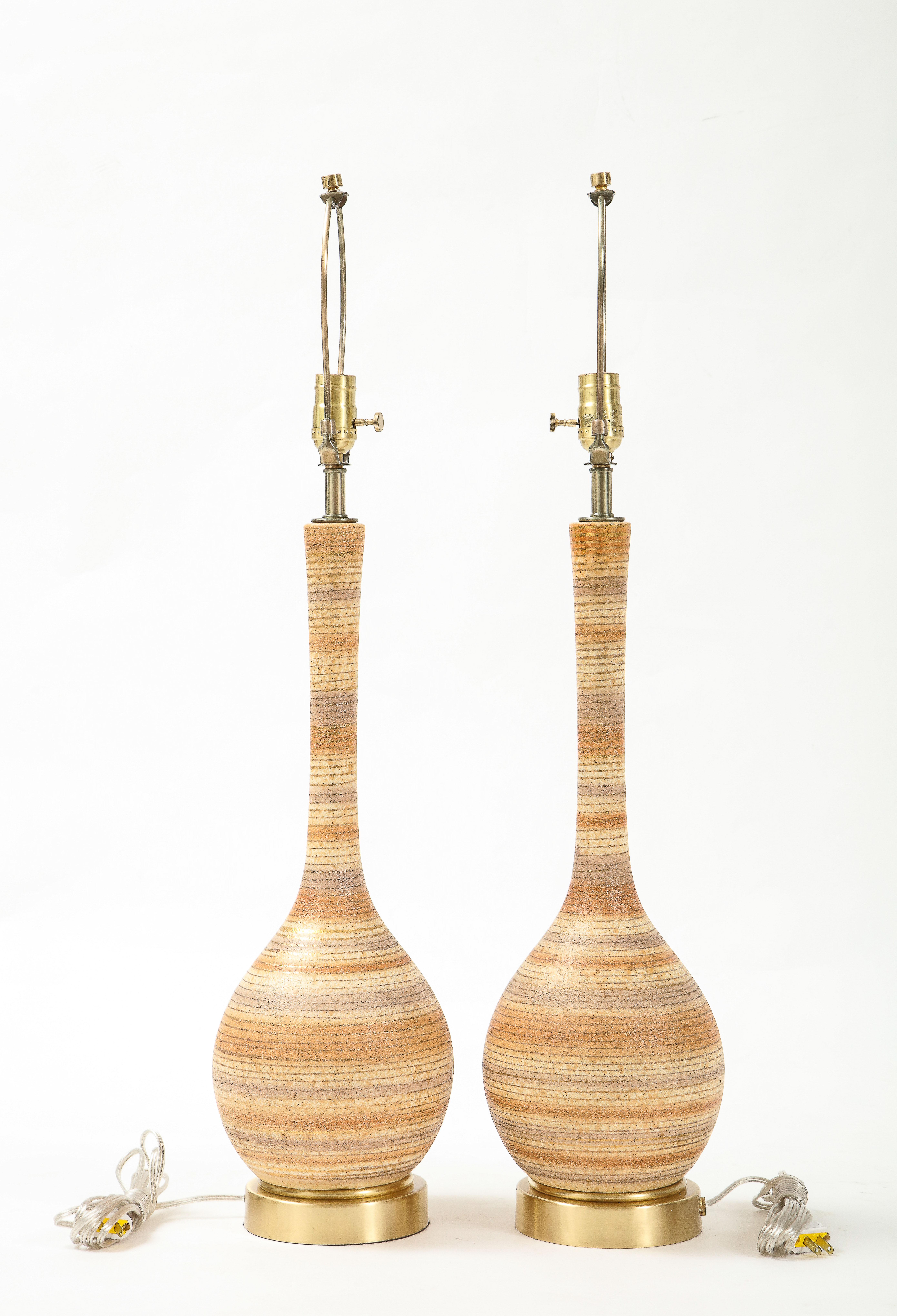 Pair of mid century ceramic lamps featuring muted tones of beige, gray and tan with a gold metallic stripe. Lamps sit on brushed brass disc bases. Rewired for use in the USA, 100W max.