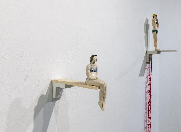 This wall-mounted figure is created with hand-built porcelain and glazes, and depicts a swimmer at the edge of a diving board.  The artist, Sascha Mallon crocheted a long red ladder that meet to the figure, but not to the ground, thus the