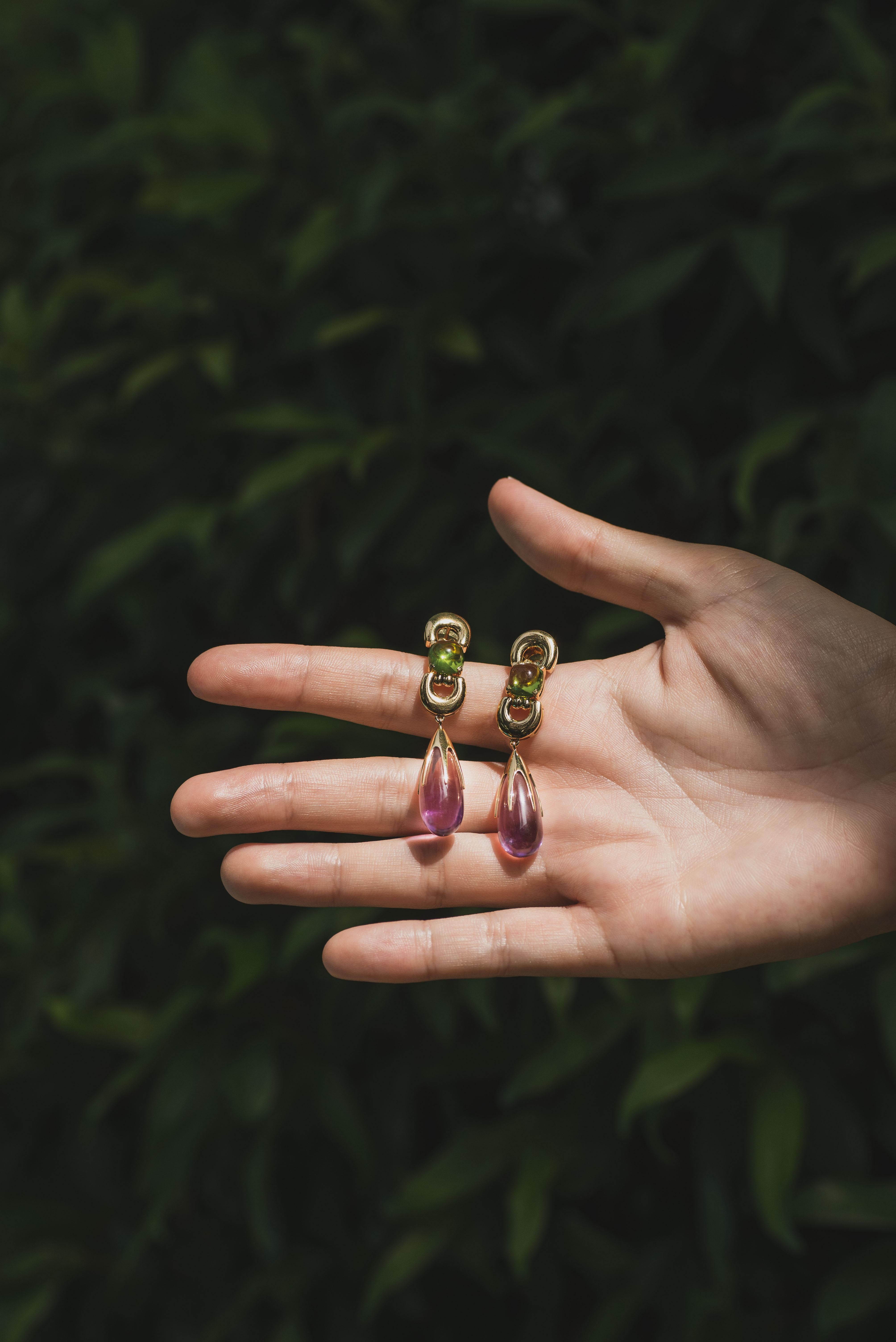 Experience the allure of Sassi Fine Jewellery's Secret of Berenice collection through these regal drop earrings. Meticulously crafted from 18k yellow gold, they feature a captivating Amethyst drop of utmost purity and exquisite cushion-cut Peridots