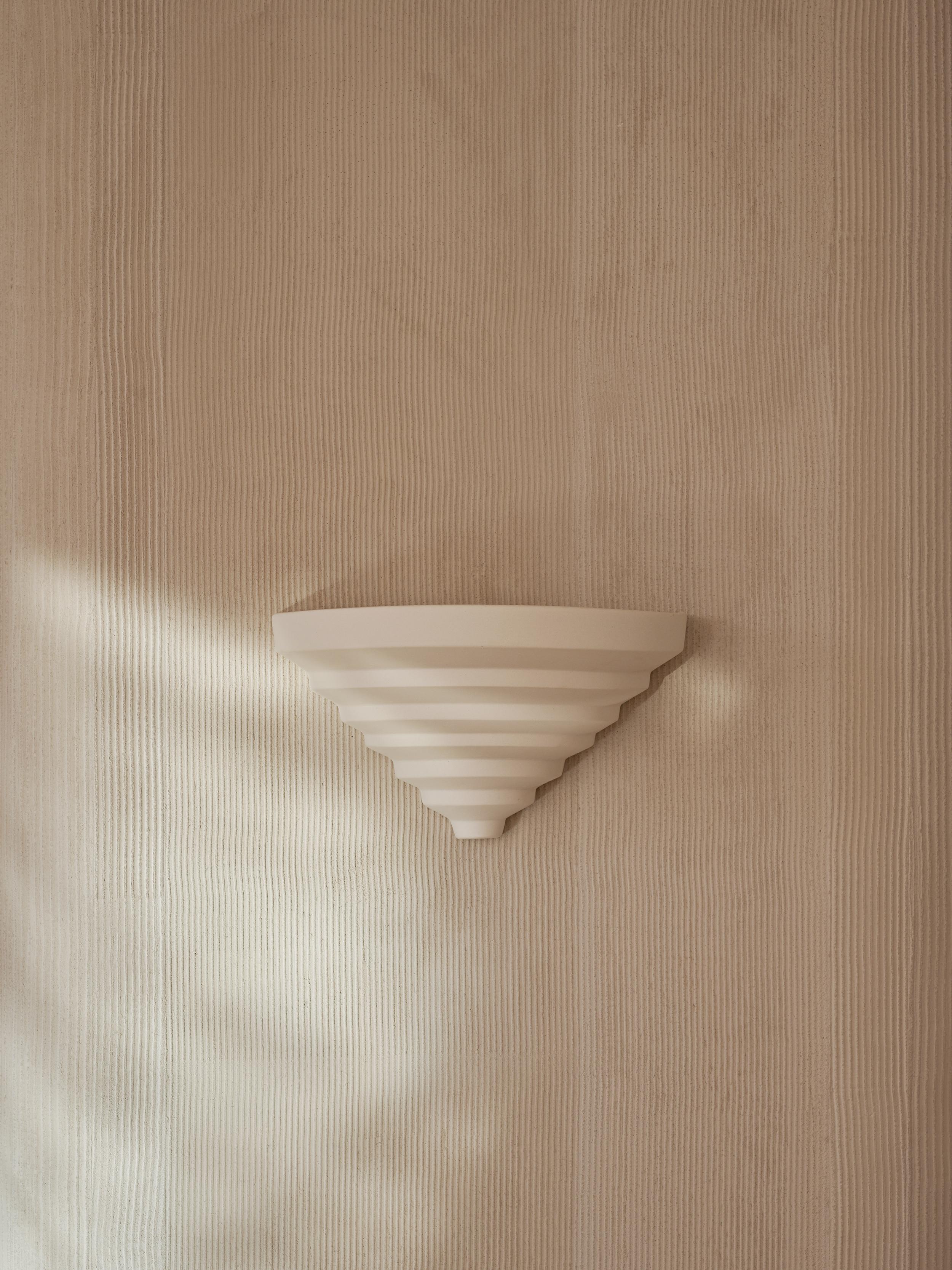 El aplique Khora Satinado combina una geometría estilizada con la calidez atemporal de la porcelana mate. Crestas geométricas que se estrechan en una elegante forma cónica.  El suave resplandor ascendente de la luz emitida lo hace ideal tanto para