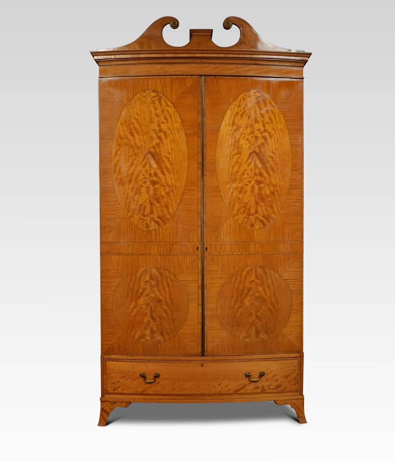Satinwood two-door wardrobe with raised swan neck pediment above two well figured panelled doors, opening to reveal large hanging area. The base section fitted with one long drawer with brass handles. All raised up on bracket feet. The wardrobe can