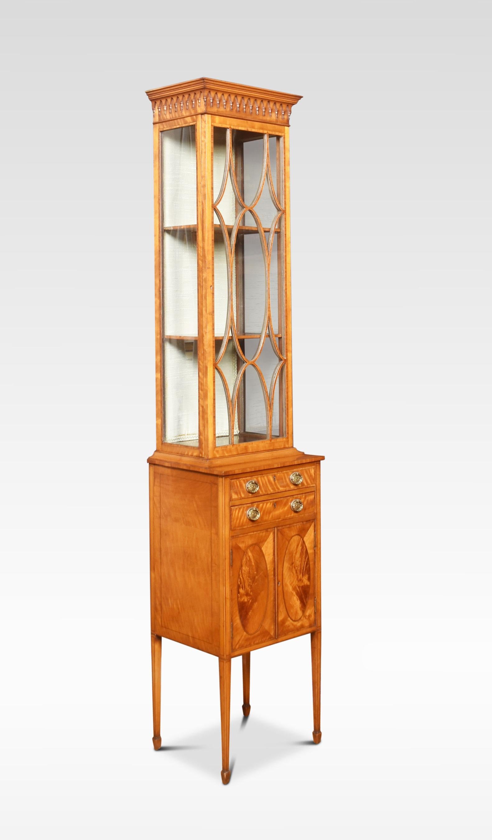 Satinwood display cabinet of small proportions, the molded cornice above carved freeze, to the large rectangular glazed door opening to reveal upholstered shelved interior. The base has a short drawer and brass tooled handles. Above a pair of