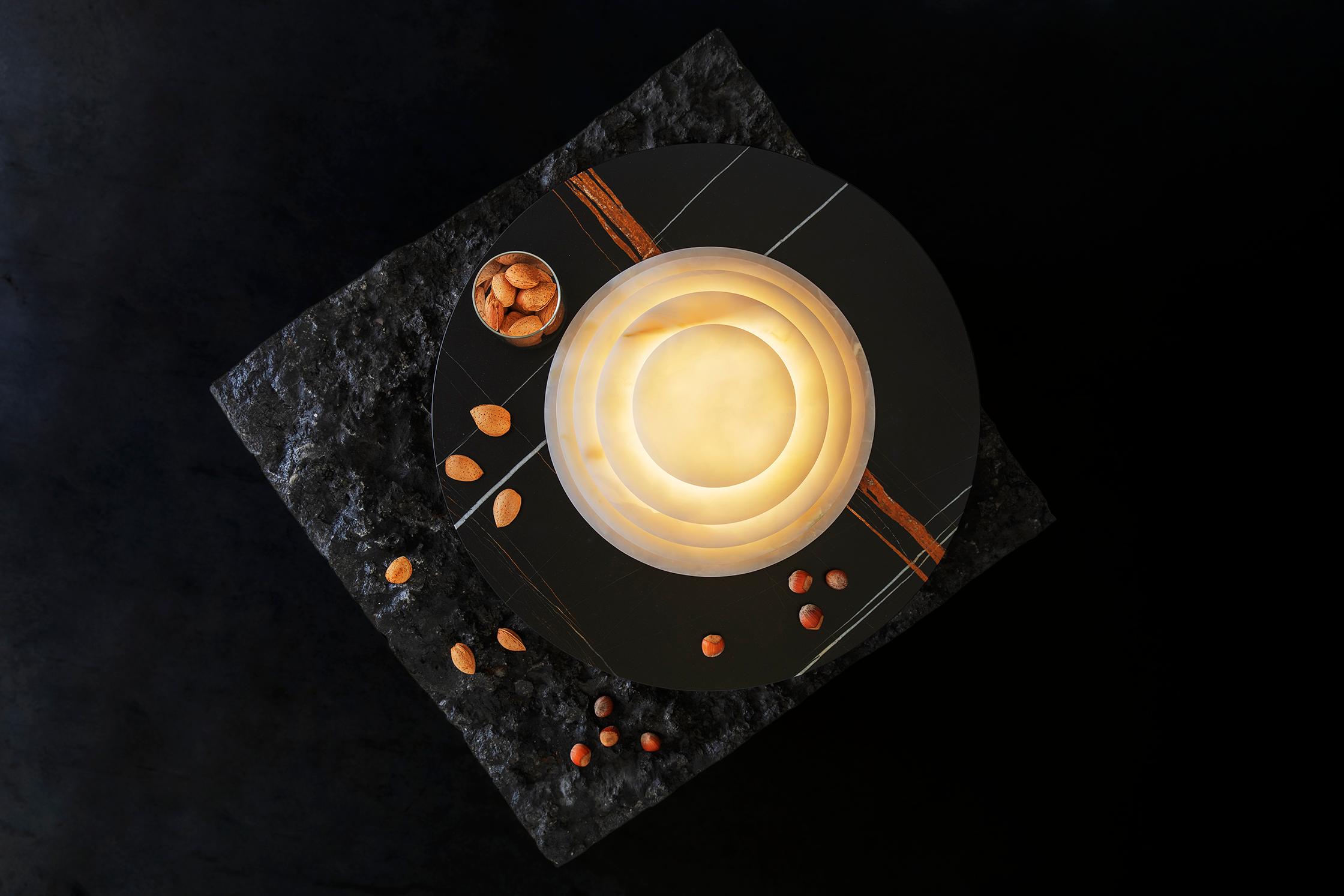 La forma circular de la lámpara recuerda la elegancia de los anillos de Saturno, creando un impacto visual único y refinado. La presencia de la bandeja, que también sirve de soporte para la apertura, hace de la lámpara un elemento de convivencia