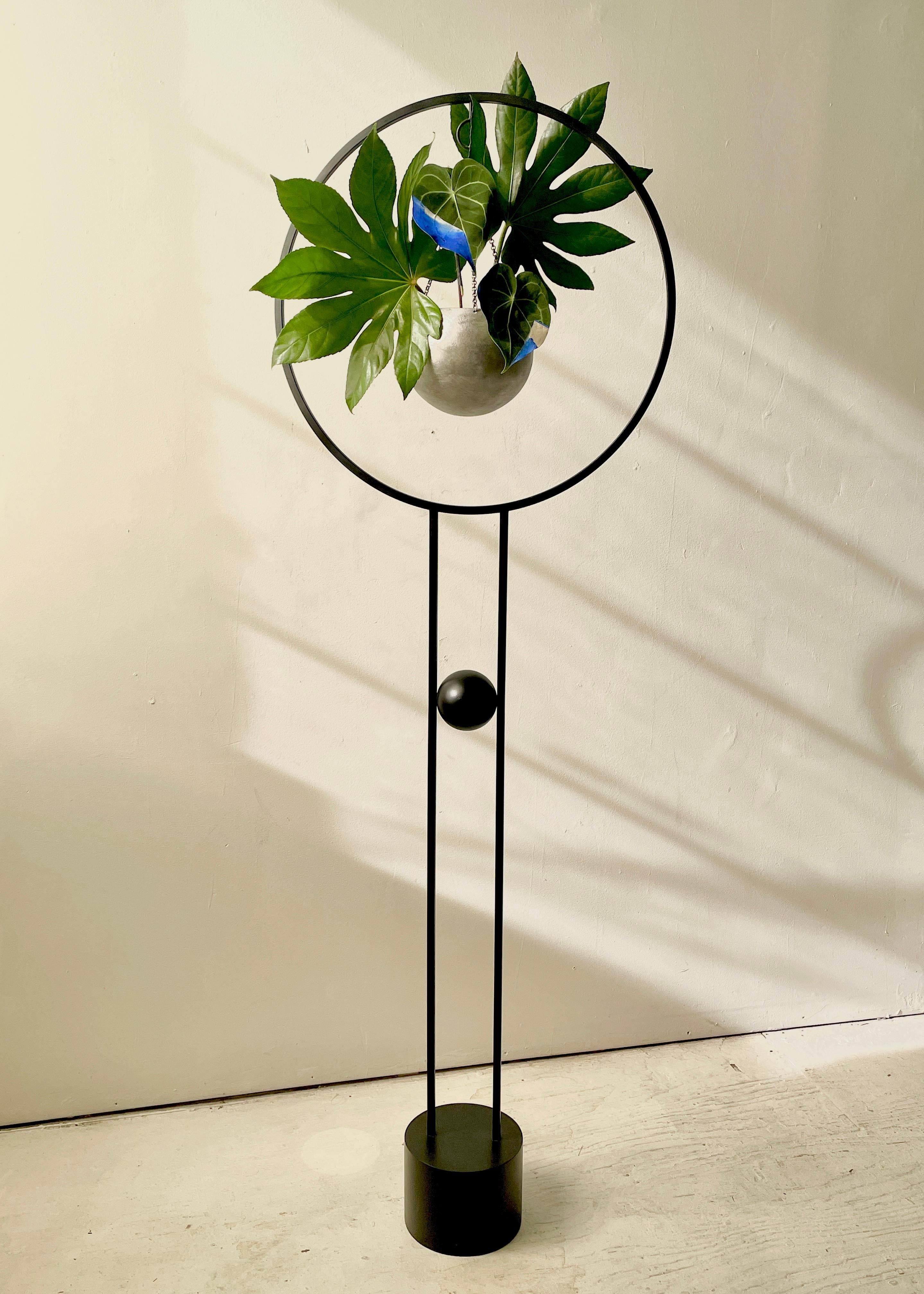Der Saturn's Return Plant Stand besteht aus einer geschliffenen Stahlkugel, die an einer zarten, aber stabilen Kette in einem massiven Stahlring hängt.
Eine kleinere Stahlkugel schwebt mühelos zwischen den beiden vertikalen Beinen, die auf einem