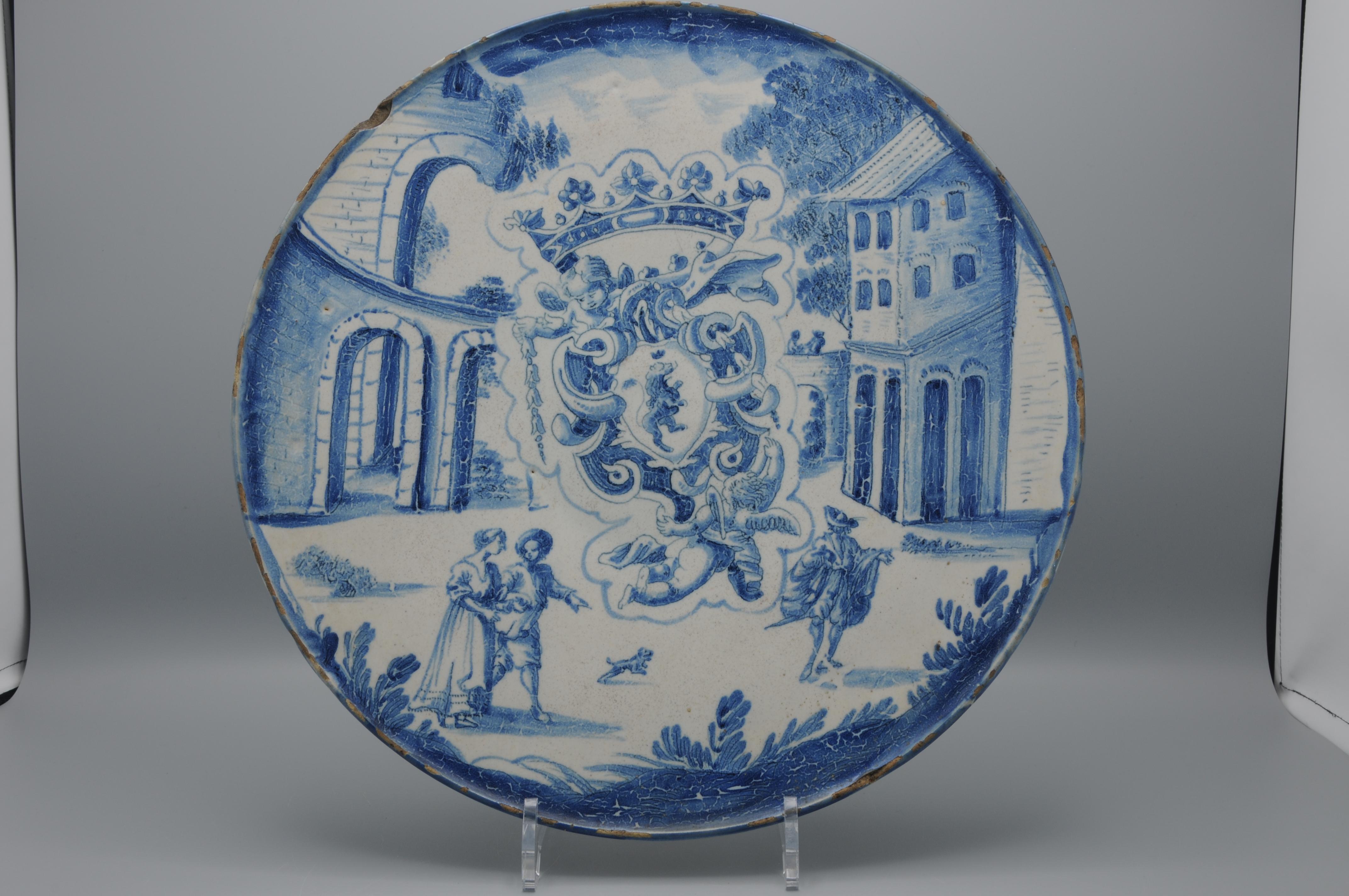 Una alzatina o crespina de Savona, plato sobre peana, con decoración azul. En el anverso un escudo heráldico con un león adornado con corona y putto. Al fondo, arquitectura elaborada con personas. 

 