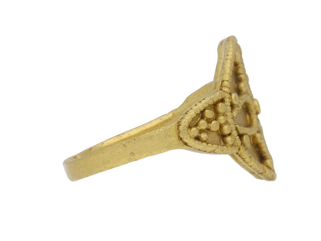 Anneau saxon en or orné. Au centre, le disque circulaire est orné d'un cruciforme ouvert, de granulations décoratives et de bordures en relief avec des détails nervurés. Les épaules en trompette sont garnies de bordures nervurées en relief et