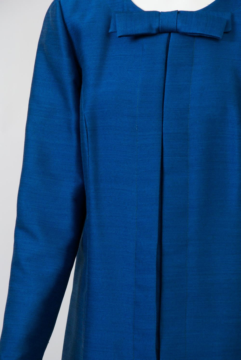 Coat-style dress of royal blue silk twill by Arnold Scaasi for the manufacturer Martini. Straight style featuring a center pleat that conceals zipper opening, low side pockets, and a fixed bow at the center of the round neckline. Long, set-in