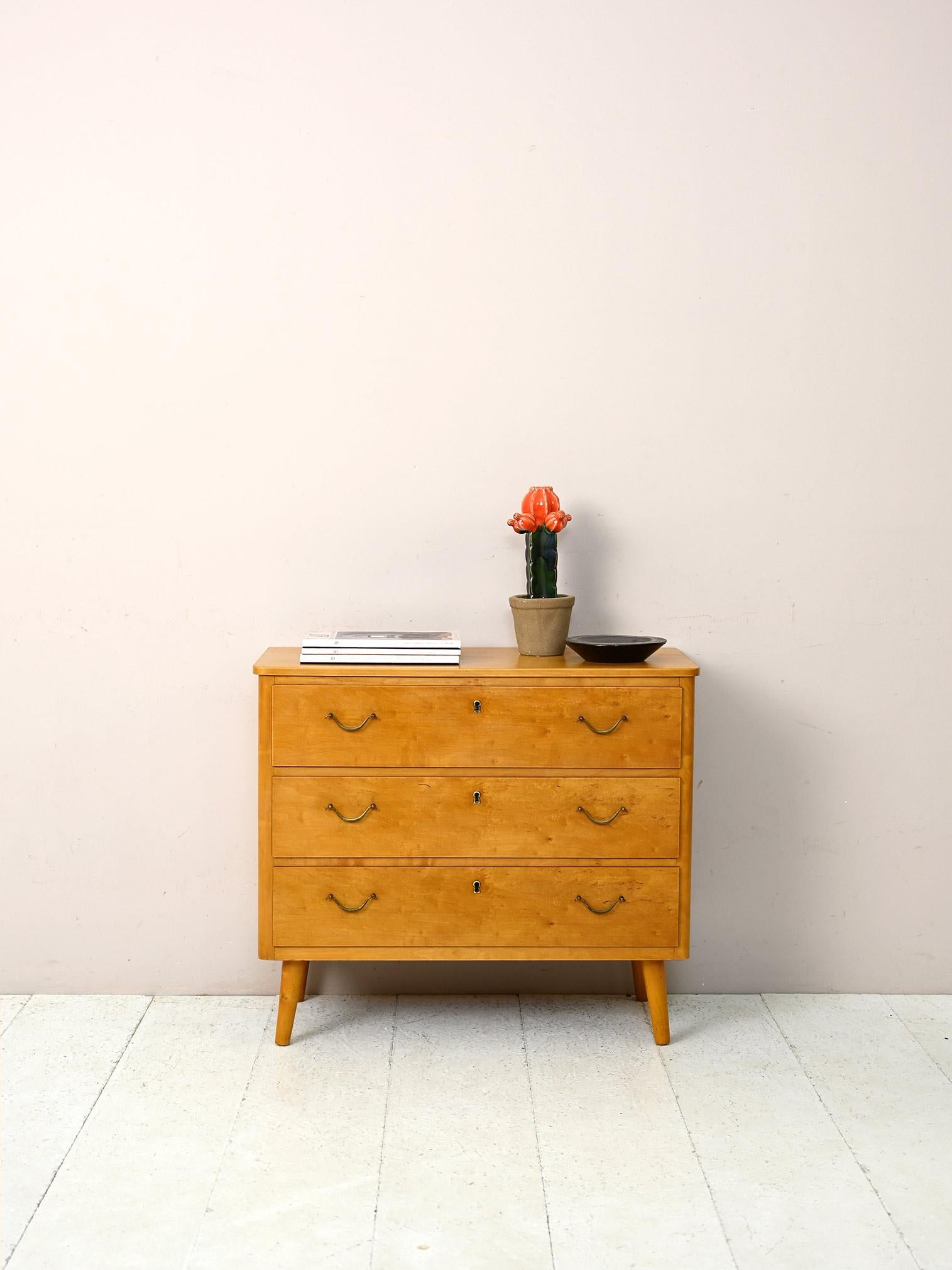 Vintage cabinet with three drawers and brass handles.

A piece of modern Scandinavian furniture with a retro feel.
The special burl wood, with its golden color and marked grain, makes this chest of drawers unique and original.
The detail of the