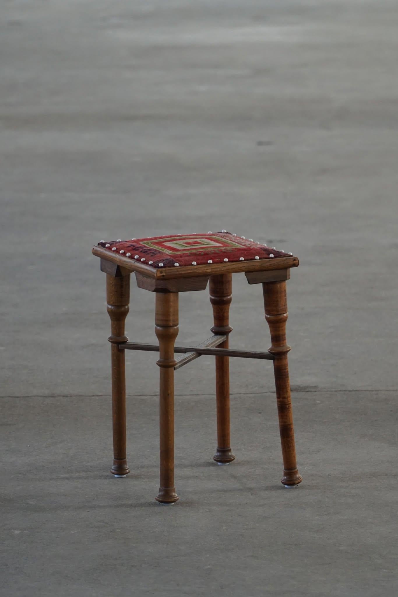 A charming little wooden stool with a square seat and four turned legs, each featuring subtle decorative shaping. The seat is covered with a patterned woven textile, showcasing a rectangular motif in red, green, and white hues. The fabric is