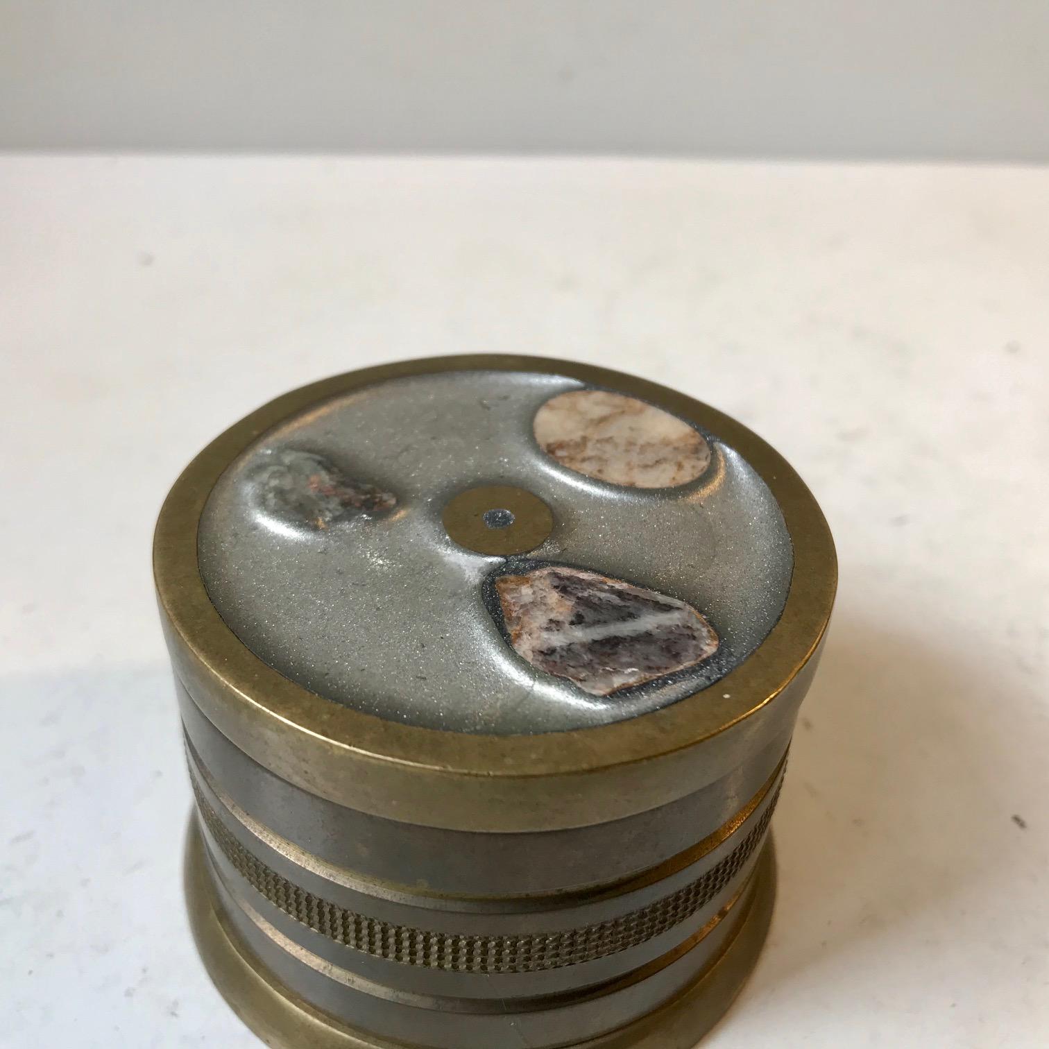 Cylindrical small trinket box fashioned from bronze. It features a removable lid covered with enamel and 3 samples of cut natural Scandinavian minerals. It was by an anonymous gold/silversmith during the 1960s. Its probably either Norwegian or