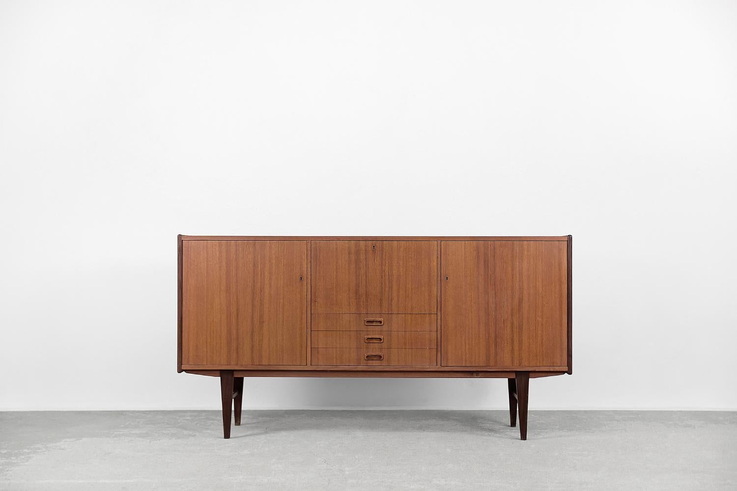 This sideboard was made in Sweden, probably in 1964. It is finished with teak wood with a warm color and regular graining. It has lockable cabinets and practical shelves. In the central part of the furniture is a vertically opening cabinet and