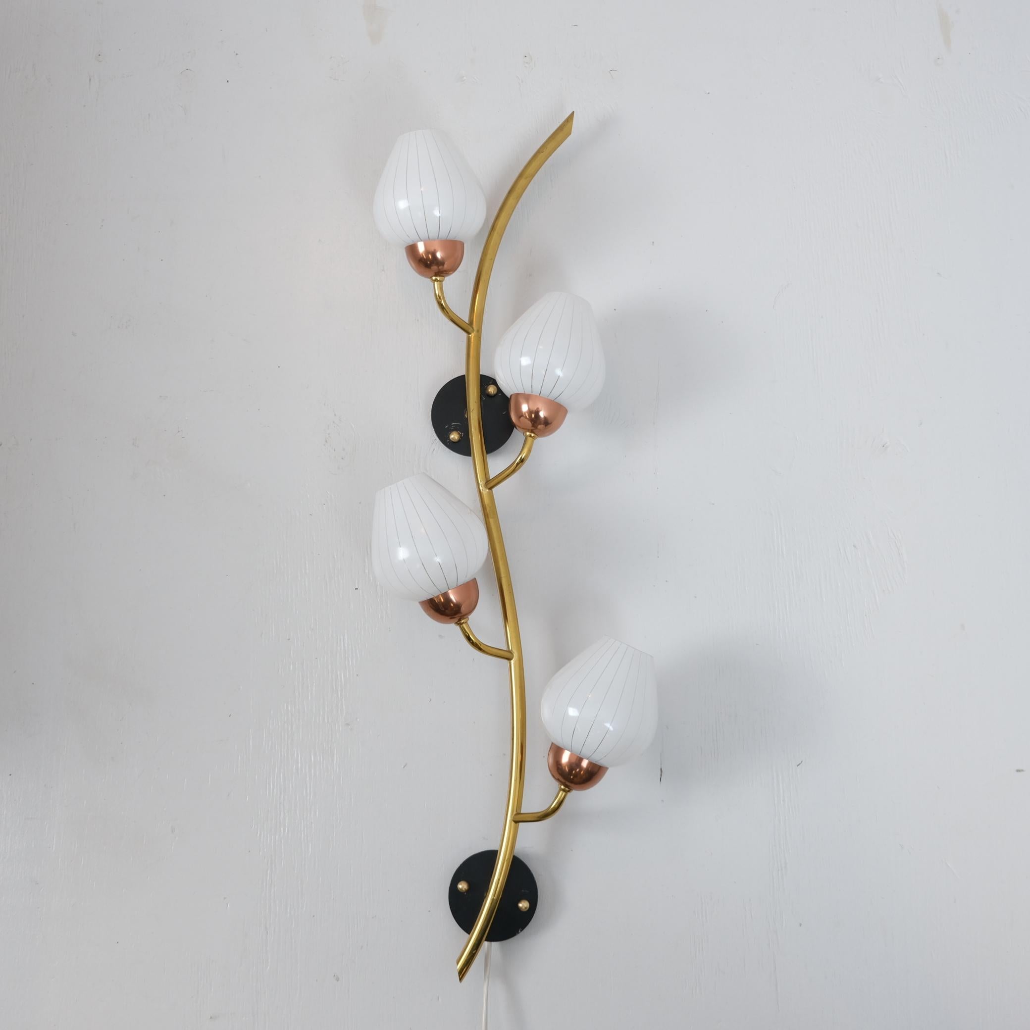 Una bellissima applique floreale con un tubo di ottone graziosamente curvato a forma di ramo e quattro delicati boccioli di rame. Ogni bocciolo è sormontato da un paralume a forma di tulipano in vetro bianco a righe. L'intero gruppo è montato su due