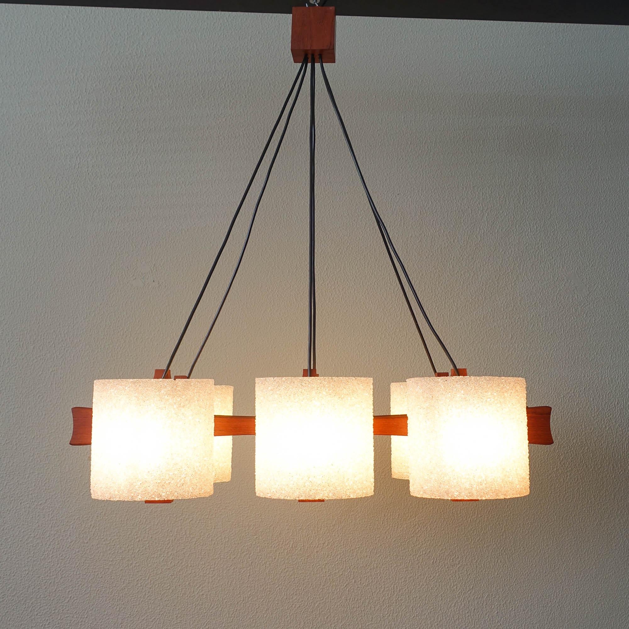 This pendant lamp was designed and produced in Denmark during the 1960's. It features a solid teak wood mount and six shades made of transparent resin granules, that due to various fused layers become opaque and gives an unique light effect. 
The