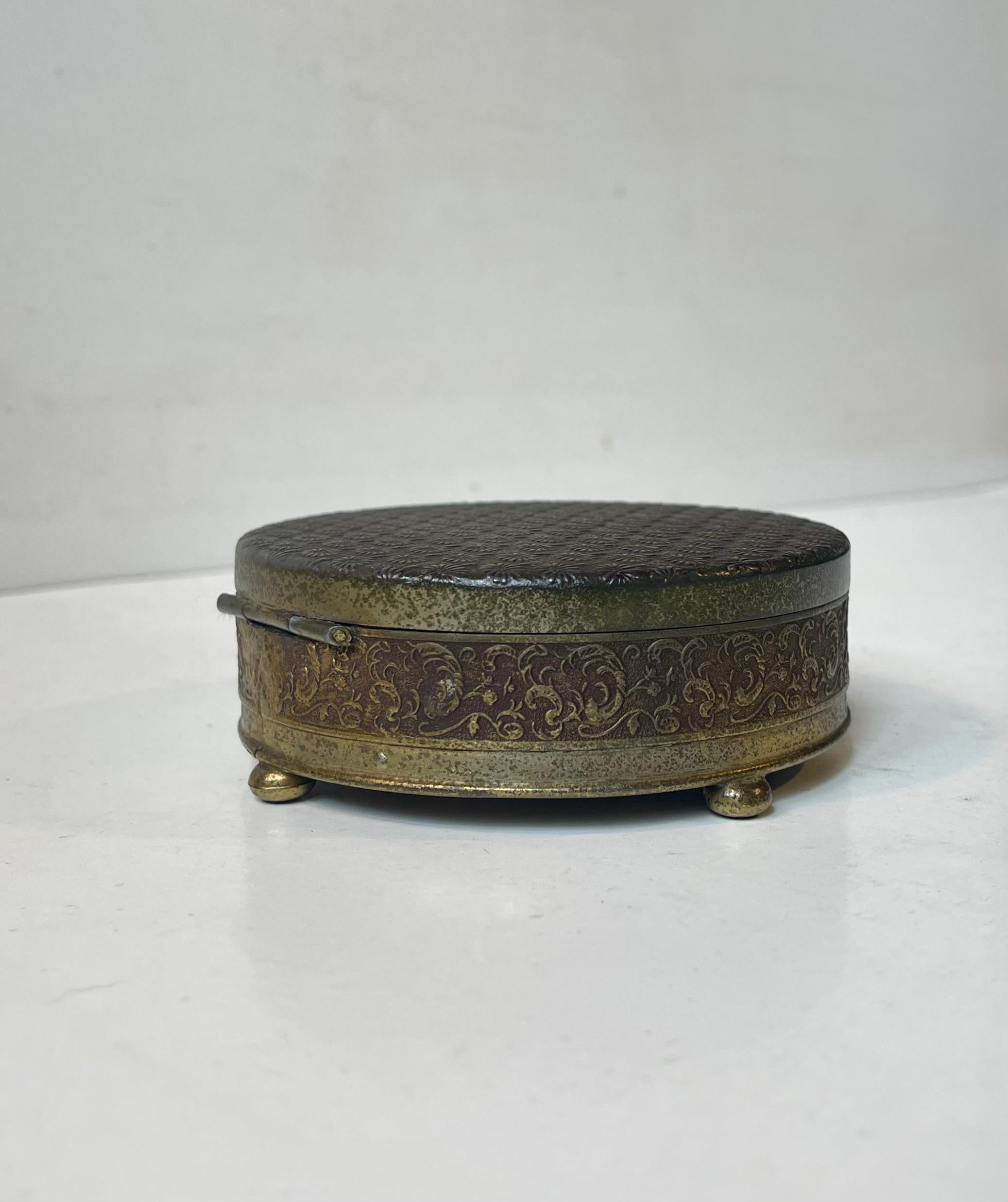 Petit bijou insolite en bronze, laiton et métal. Le couvercle et le fond sont décorés de marguerites. Son intérieur est tapissé de textile vert. Elle a été fabriquée à la fin des années 10 ou au début des années 20 par un bijoutier anonyme,