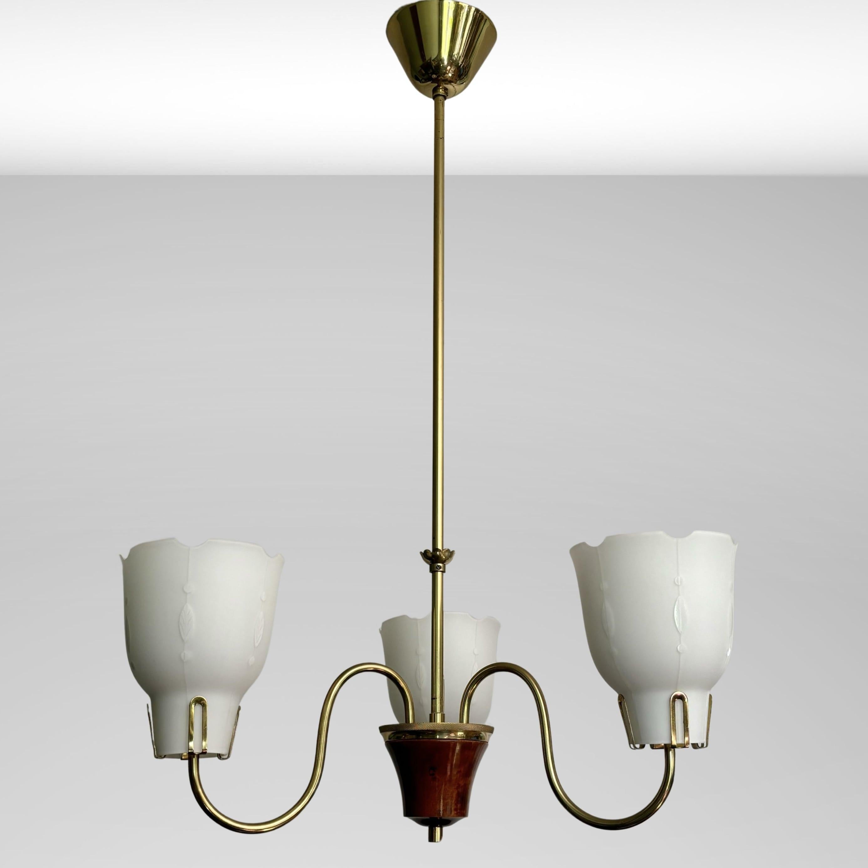 A Swedish 1940s chandelier made from brass and walnut with three curved arms holding bell-shaped shades in opal glass. Each shade has a discreet white leaf pattern, adding an extra touch of elegance. With its minimalistic shape and clean lines, this