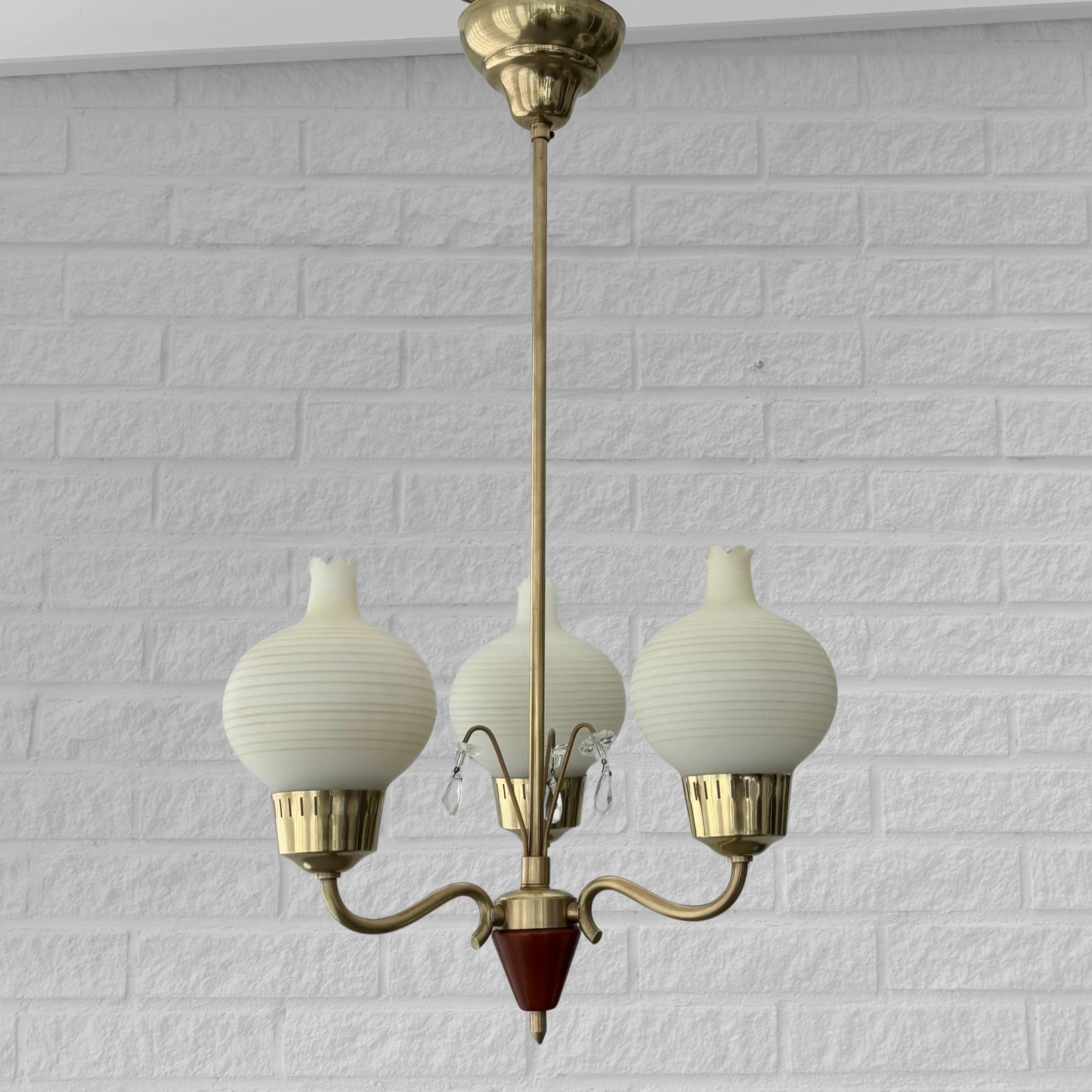 A Scandinavian modernist chandelier crafted from brass, featuring three curved arms with a wooden cone at the base and decorative crystal glass prisms at the center. The fixture is fitted with three beautiful glass shades adorned with subtle stripes