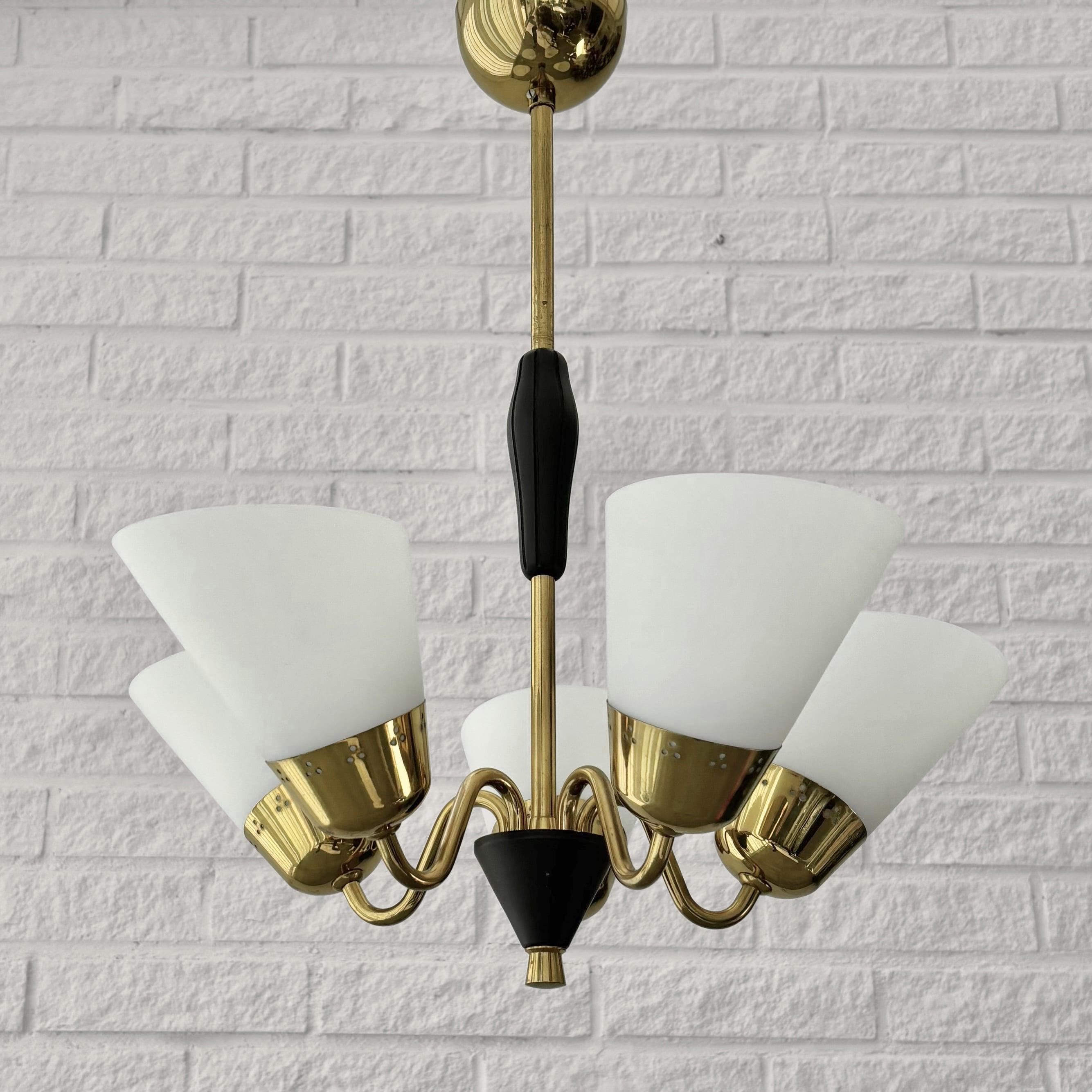 A Scandinavian modernist chandelier crafted from brass, featuring five curved arms supporting conical opal glass shades. The shades are secured by brass fittings perforated with a decorative dot pattern. The fixture also includes a black wooden