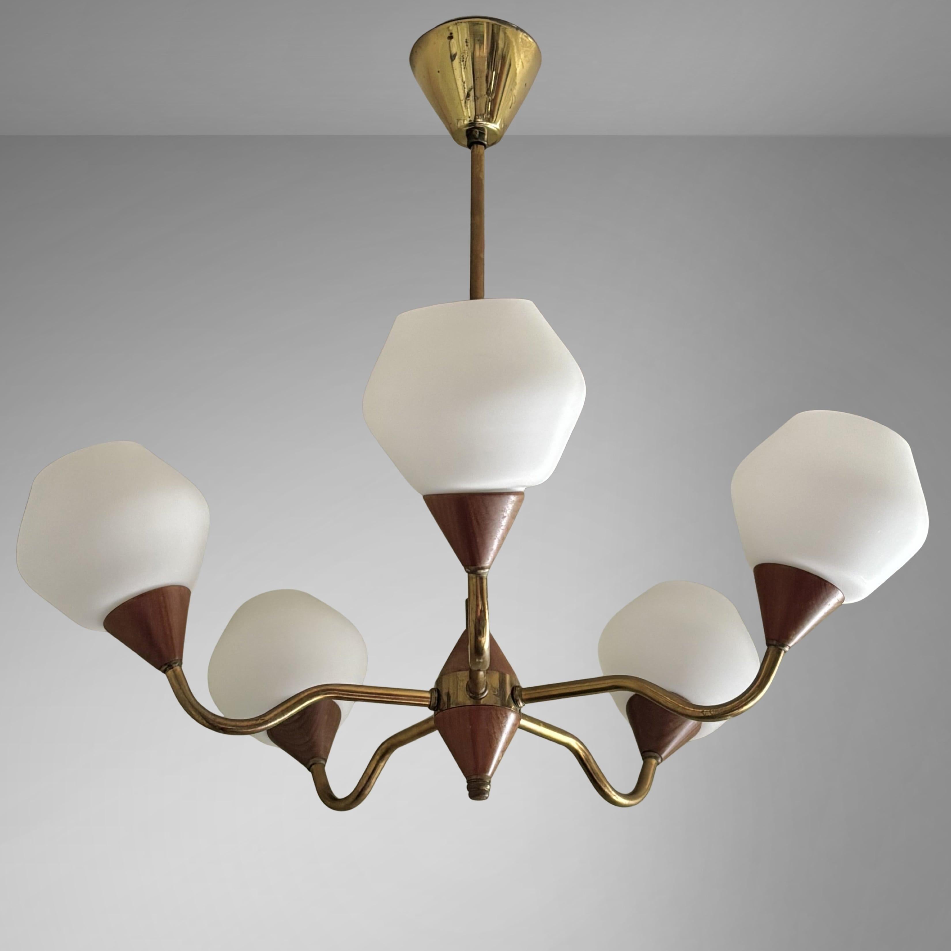 Scandinavian mid-century chandelier, produced by an unidentified Swedish manufacturer in the 1950s. Crafted from brass, it features five curved arms extending from a cone-shaped centerpiece, each holding a bell-shaped white glass shade. Complete