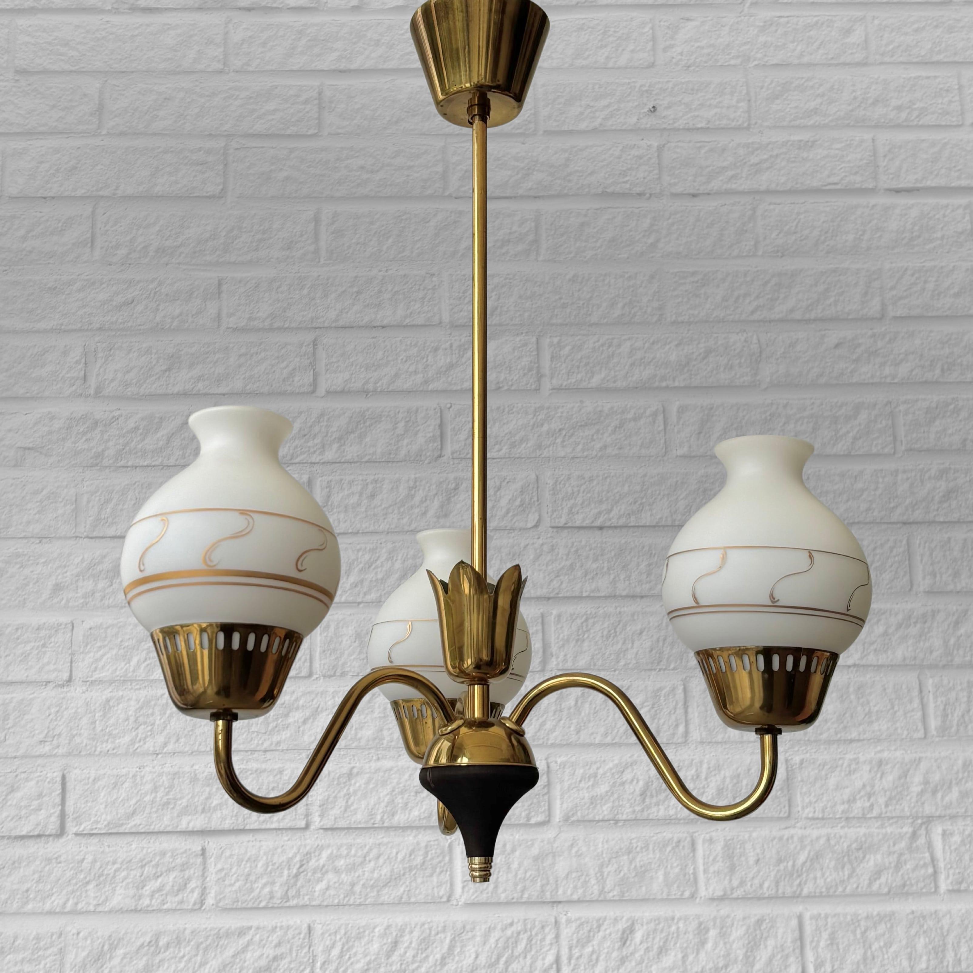 Araña modernista escandinava de latón, con tres brazos curvos y un cono de madera en la base. La luminaria está equipada con tres hermosas pantallas de cristal adornadas con un motivo dorado pintado a mano. Incluye el dosel de techo original de