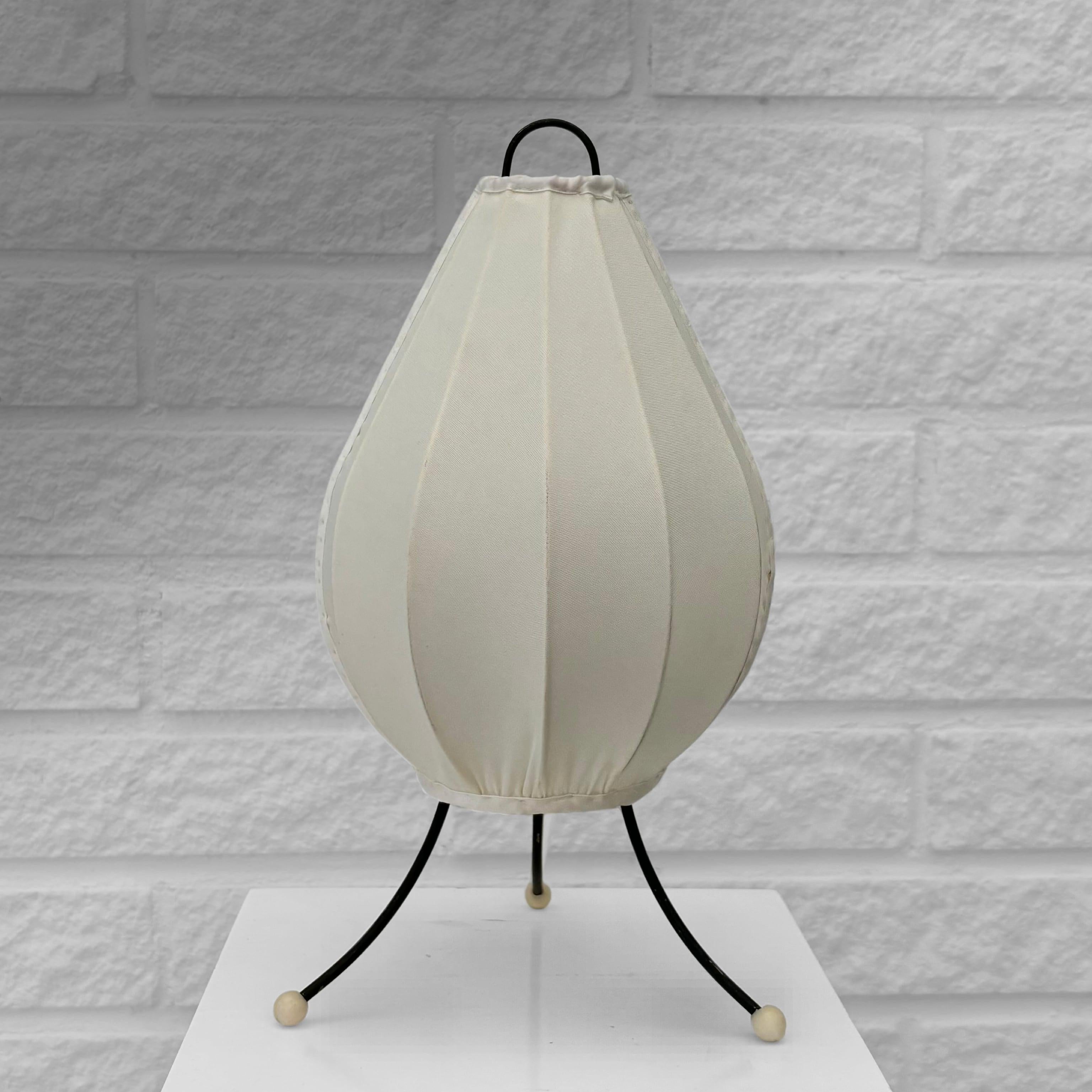 A modernist table lamp inspired by traditional Chinese lanterns, crafted from black metal strings with brass details. It features three thin metal legs with small plastic feet. The drop-shaped shade is covered in a silk-like textile, giving the