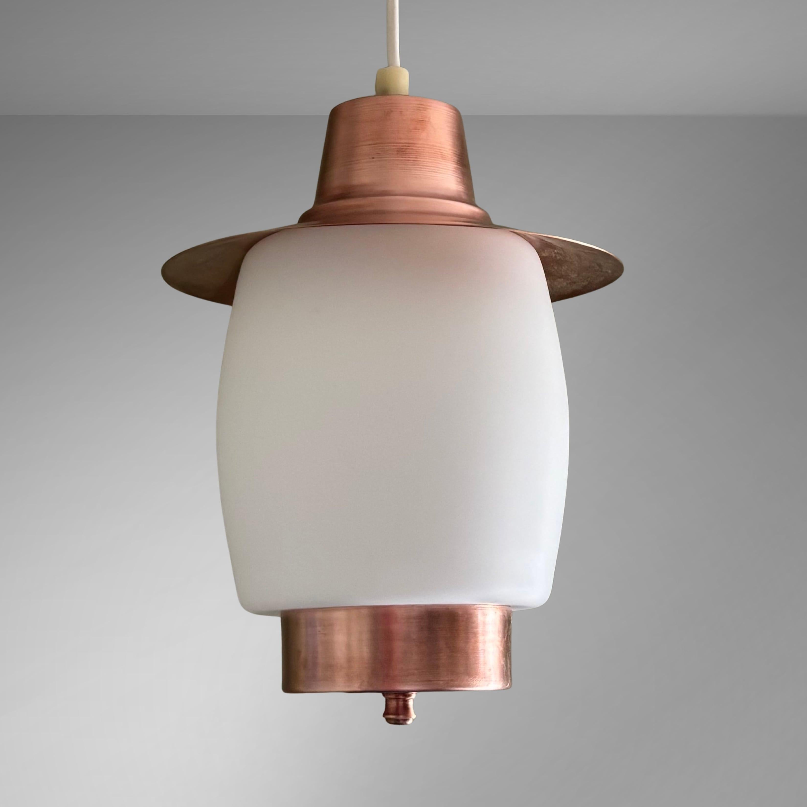 A minimalist mid-century pendant lamp, produced by an unidentified Swedish manufacturer in the 1950s. It is constructed with a disc-shaped upper shade and a bowl-shaped base, both crafted from solid copper, with a rounded opal glass shade in