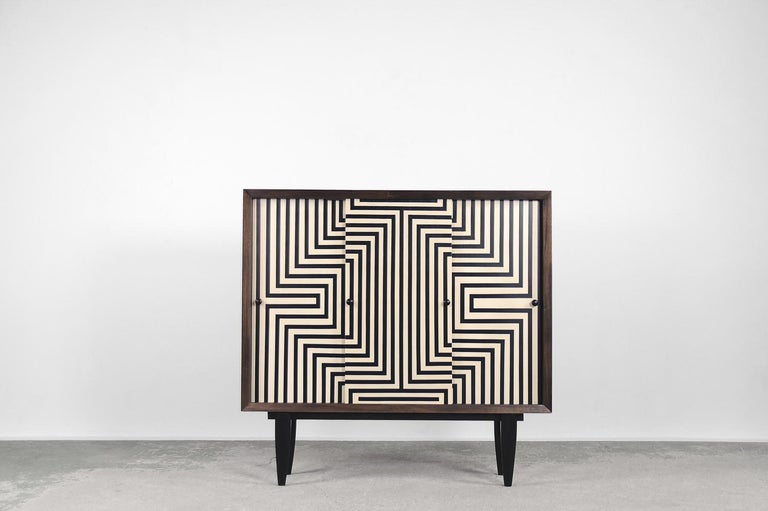 This cabinet was made in Scandinavia during the 1960s. This piece was finished in birch wood with interesting graining and tones in Earth colours. It has three sliding doors decorated with hand-painted, black and white, linear painting in the Op-Art