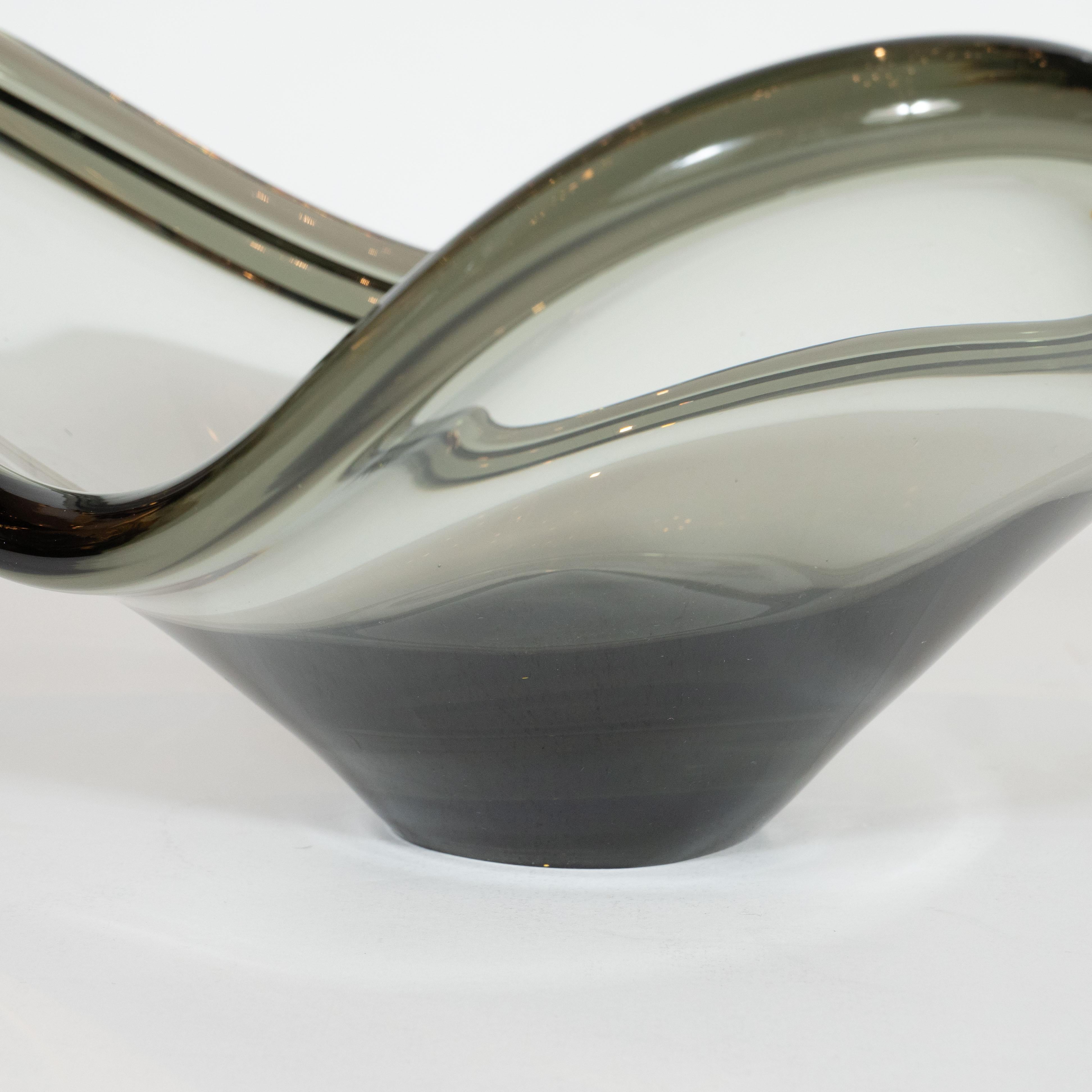 This refined and sculptural Mid-Century Modern bowl was hand blown by the celebrated Swedish maker Holmgaard in 1960. It features a sculptural three sided atomic form with undulating sides and a subtly raised lip all realized in a translucent smoked